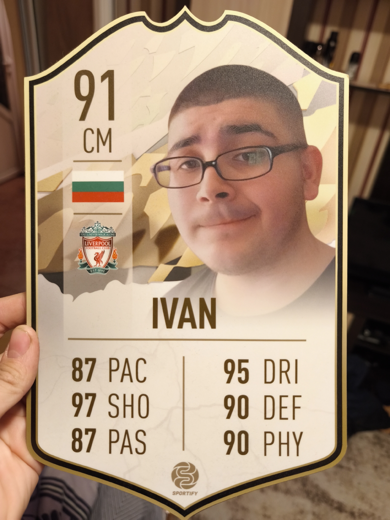A person holds a custom-made card resembling a FIFA Ultimate Team (FUT) card. The card features the holder’s photo, set against a gold-toned background, with the number "91" and the unit "CM" displayed at the top left. Beneath these, there's the flag of Bulgaria, the country the cardholder represents. Below that is the logo of Liverpool Football Club, which might indicate the cardholder's allegiance or team affiliation. The name "IVAN" is displayed prominently below the picture. Underneath the name, the card lists football-related statistics, including "87 PAC," "97 SHO," "87 PAS," "95 DRI," "90 DEF," and "90 PHY." The "SPORTIFY" logo is located at the bottom center. The location is not specified but the flag indicates the cardholder is from Bulgaria. No further information indicates the city of Burgas. The holder's hand is slightly visible at the left. The background is somewhat blurred, showing parts of a room with furniture.