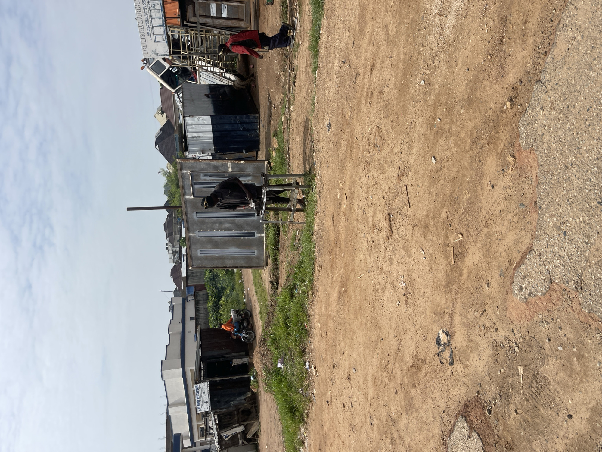 The image depicts an outdoor scene in what appears to be a workshop or scrap yard, with a dusty, dirt ground and some paved areas. The scene is oriented vertically, suggesting the photo was taken with the camera rotated.
In the foreground, a person wearing a dark shirt and pants is seated at a table made of metal, holding papers and appearing to be working. Behind them, a metal structure with corrugated metal sheeting serves as a shelter or part of a building. Further back, several metal structures and containers are visible, some appearing to be workshops or storage units. One building has a sign with text that reads "AR NEATWORKS METAL GATES, WINDOWS, WELDING, ALUMINIUM, BORROW & DOORS."
Another person, wearing a red jacket and dark pants, is standing further back near one of the structures, looking downwards. There is also a motorcycle parked near a building, with a person wearing an orange shirt on it. The environment suggests a somewhat utilitarian and possibly busy setting.
The sky is mostly overcast with pale clouds. The lighting suggests it might be daytime, perhaps late morning or afternoon. The overall atmosphere seems to be one of industrial activity and manual labor. There are some green patches of grass and vegetation interspersed between the structures. The ground is mostly dry and dusty, with some debris scattered around.