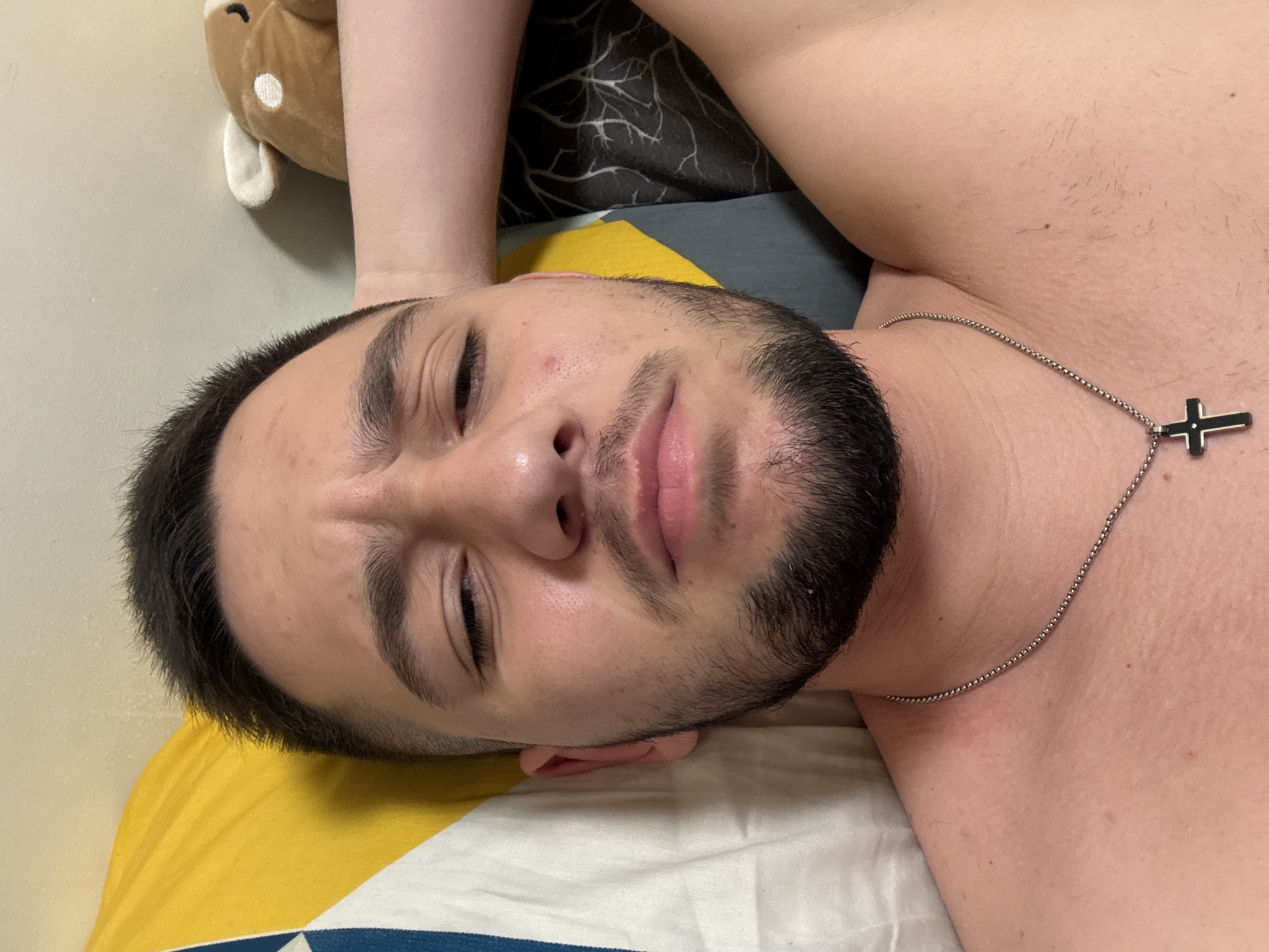 A close-up, medium-shot image features a man lying on what appears to be a bed. His head rests on a yellow and white patterned pillow. His face is centered and fills a large portion of the frame. He has short dark hair, a beard, and his eyes are partially closed. He wears a silver chain necklace with a black cross pendant. The background includes a brown stuffed animal and a dark patterned bedspread. There is nothing in the image to indicate the location is Sofia, Bulgaria.