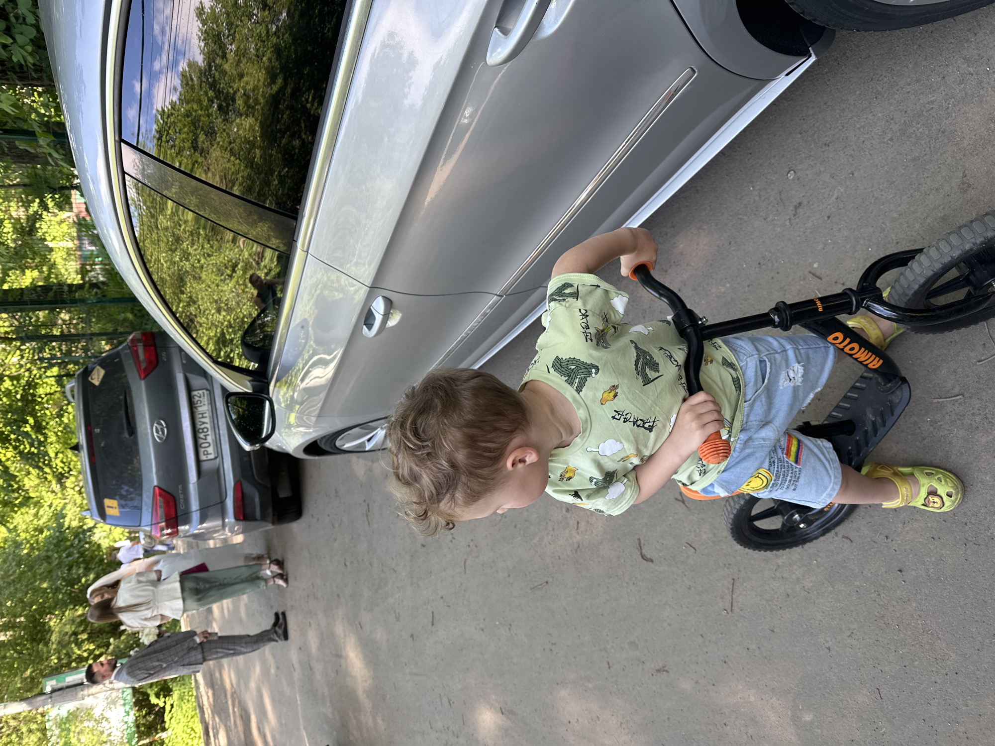 The image, taken outdoors in daylight, is oriented sideways and features a young child on a balance bike next to a parked car, with two adults and another car in the background.
The primary subject is a toddler, approximately 2-4 years old, with curly light brown hair, wearing a light green t-shirt patterned with cacti and clouds, light blue ripped denim shorts with a rainbow patch and yellow smiley faces, and bright yellow sandals featuring a Mickey Mouse head. The child is holding the handlebars of a black and orange balance bike, branded "micro," and appears focused on riding.
To the child's left is a shiny silver or light grey sedan. Its side reflects green trees, blue sky, and possibly a person, indicating clear weather. Further in the background, a dark grey Hyundai Creta SUV is parked. Its Russian license plate, "P048YH 152," is clearly visible, with "152" being the region code for Nizhny Novgorod.
Behind the cars and child, two adults are visible. One is a man in a grey plaid suit, white shirt, and dark tie, standing upright. The other is a woman in a white top and a long dark green skirt, holding a white bouquet, suggesting a formal event like a wedding. Lush green foliage and trees line the background, contributing to an outdoor, verdant setting. The ground is a paved surface, likely asphalt or concrete. The bright lighting and reflections suggest it is a sunny day.
Text visible includes "micro" on the balance bike, "P048YH 152" on the SUV's license plate, and partially discernible English words such as "TOTEM" and "NEW YORK" on the child's shorts.
