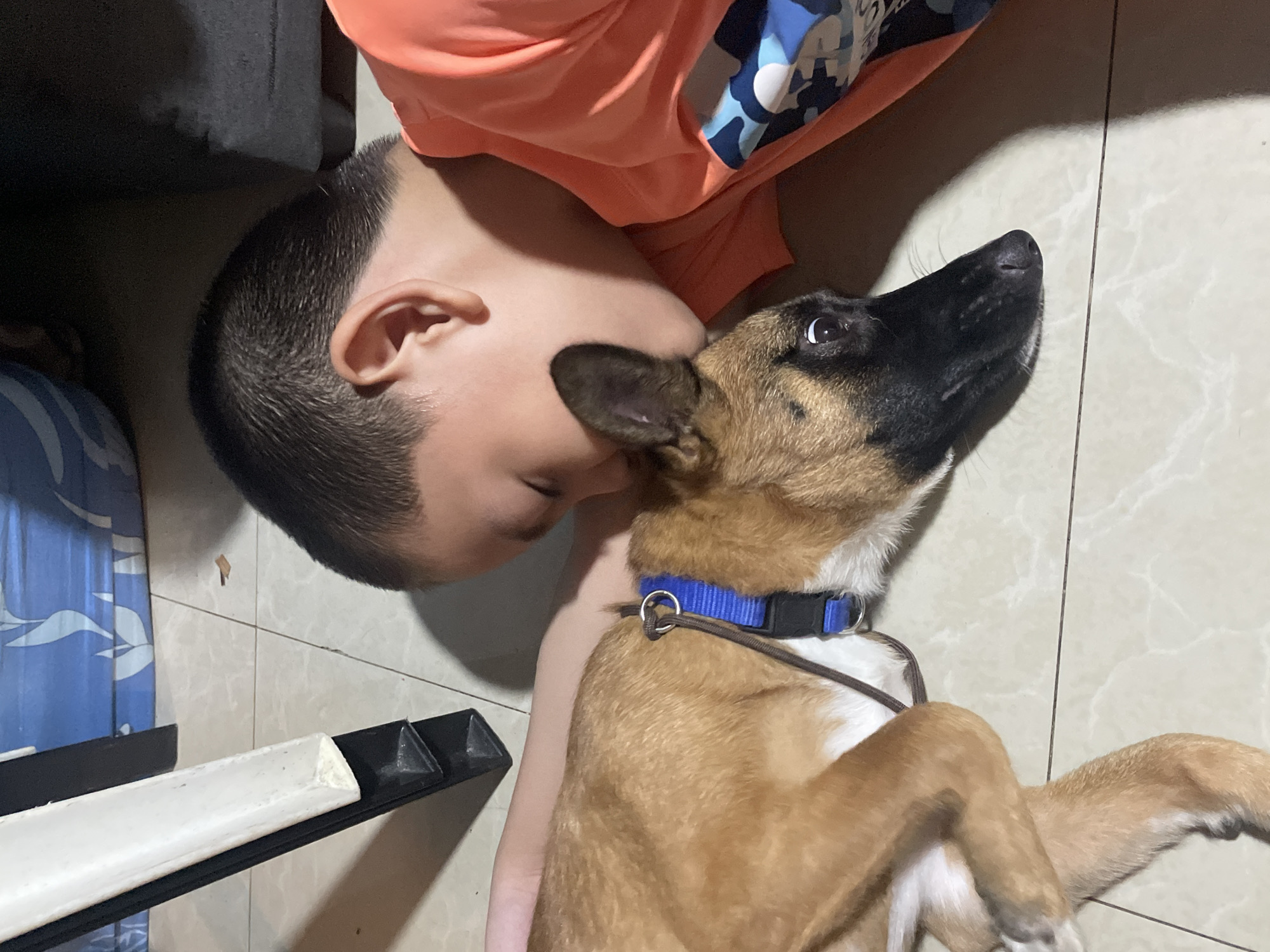 A young boy, wearing an orange shirt with a camouflage pattern on the sleeve, is lying down on a tiled floor, with his head resting on a dog. The dog, a medium-sized breed with brown and black fur, is also lying down. The boy appears to be embracing the dog. In the background, part of a white and black object, possibly furniture, is visible, along with a blue patterned fabric. The scene is dimly lit, casting shadows on the floor. The image was captured in Davao City, Philippines.
