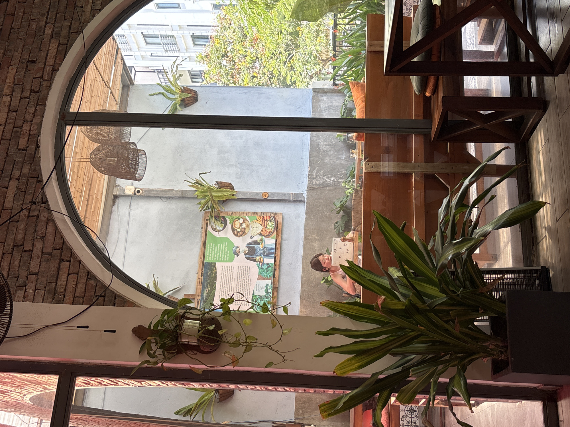 This image shows an interior view of a cafe in Da Nang, Vietnam, looking out through a large arched window. The cafe has exposed brick walls and wooden furniture. A woman is seated at a table, looking at a laptop, with a plant in a pot in the foreground. Outside the window, there are trees and buildings, suggesting an urban setting. The overall atmosphere appears to be casual and relaxed.