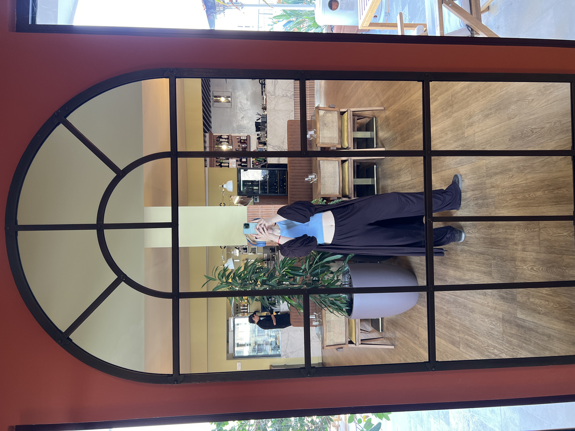 A window with a semi-circular top section reflects the interior of a building with light wood floors, tables, chairs, and a woman holding a cell phone taking a selfie. The woman is wearing a blue top and black pants. There is also a person in black standing behind a potted plant. The reflection shows a bar or counter with bottles and glasses. The window frame is dark metal. The wall around the window is painted in red. The top of the window shows an outdoor area with chairs. The building appears to be located in Boreti, Montenegro.