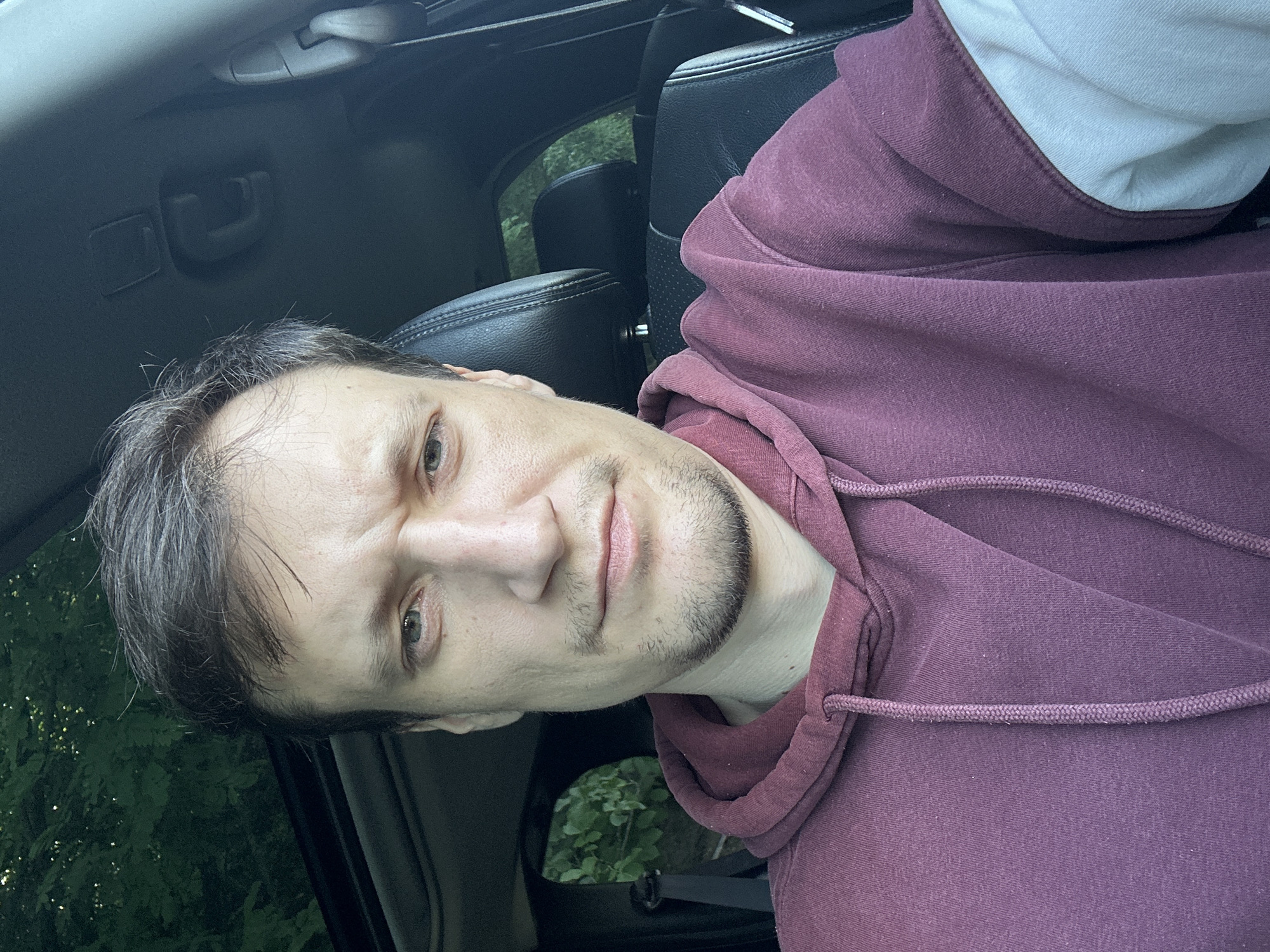 A man is sitting in the driver's seat of a car. He is wearing a maroon hoodie over a white long-sleeved shirt. The car's interior is visible, including the black leather seats and the sunroof. Trees and foliage are visible through the car windows and sunroof, suggesting an outdoor setting, likely in a wooded area. The man is looking at the camera with a neutral expression. No specific location in Makiv, Ukraine, can be identified from the image.