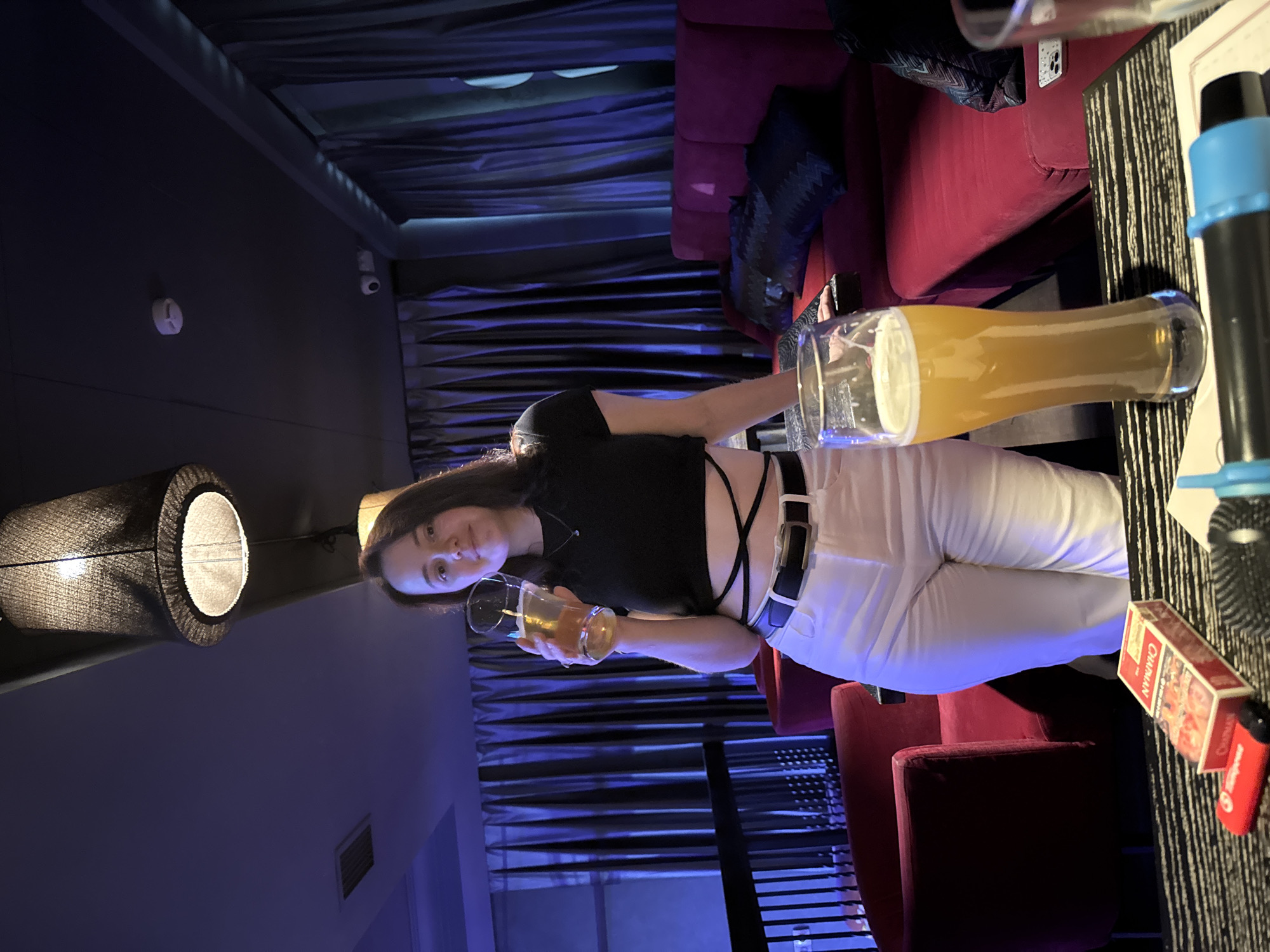 A young woman with brown hair stands in a dimly lit, possibly a bar or lounge, holding a glass of beer. She is wearing a black crop top, white high-waisted pants, and a black belt. The background features plush red seating and dark curtains. On the table in front of her are a pack of Chapman cigarettes, a red lighter, and two glasses of beer, one of which is in the foreground and appears larger. The scene is illuminated by a large pendant lamp and ambient colored lighting. There are security cameras visible on the ceiling, suggesting a public establishment. The overall atmosphere appears relaxed and social. The presence of Chapman cigarettes, a Russian brand, might suggest the location is in Russia, potentially Izhevsk.