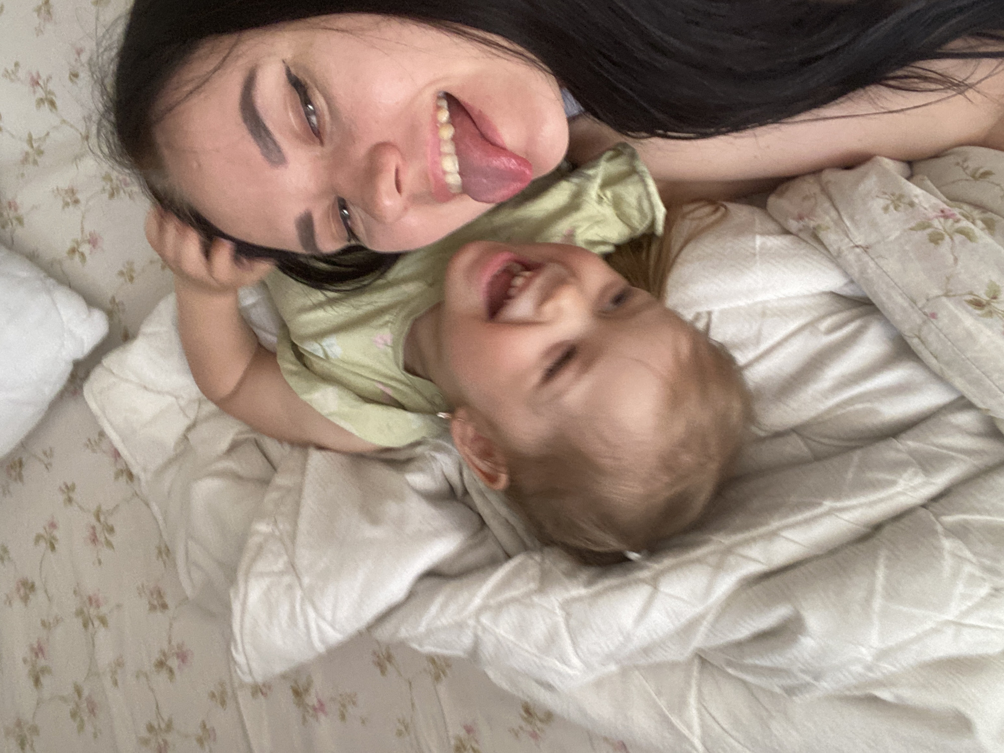 A woman with long, dark hair and exaggerated makeup lies on a bed with floral print sheets, sticking her tongue out playfully. A young child with fair hair and a green shirt is nestled next to her, laughing with their mouth open. The scene appears to be indoors on the bed, covered with a white quilt. The location is unknown.