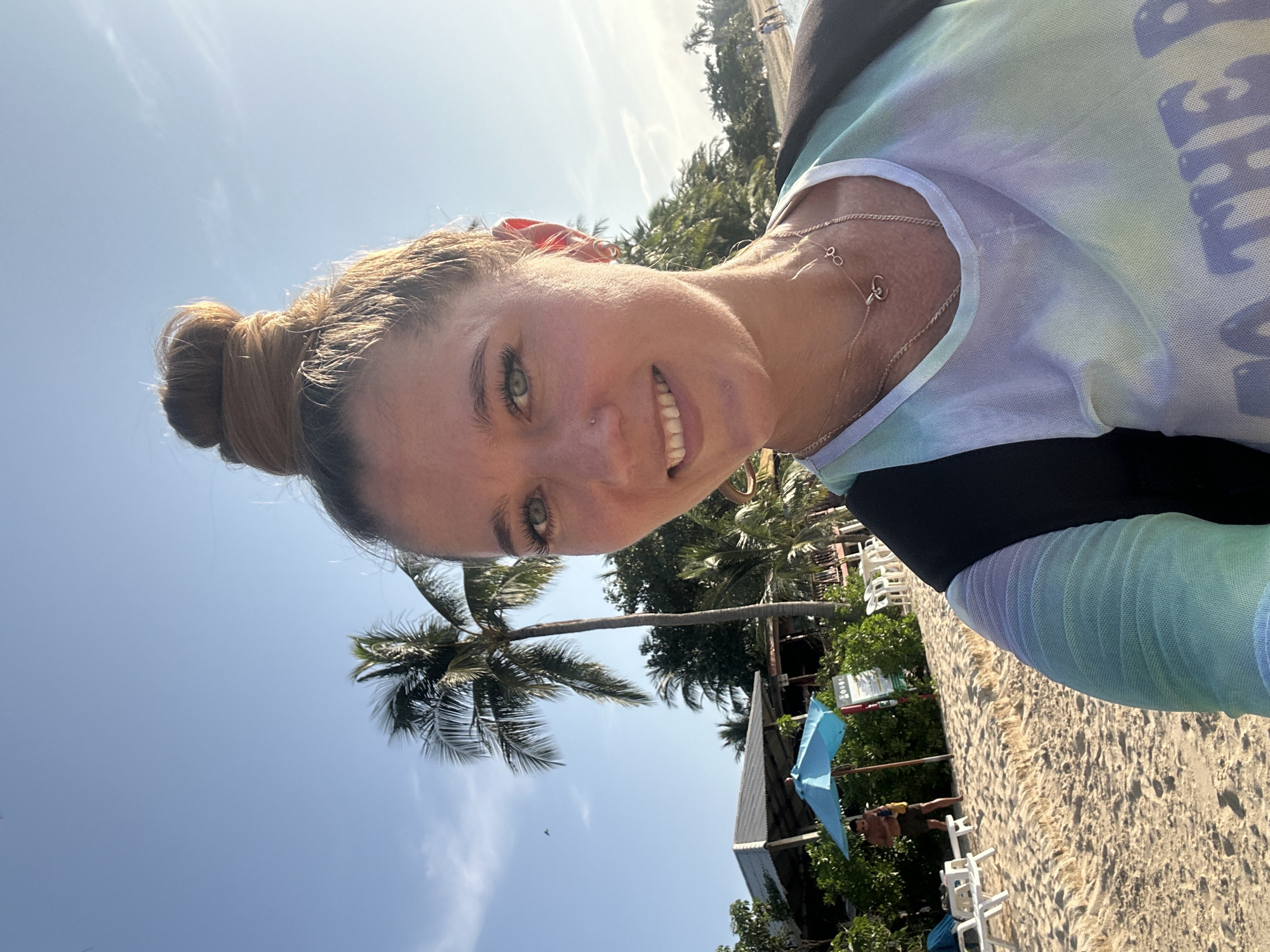 In Ko Samui District, Thailand, a young woman with blonde hair in a messy bun takes a selfie on a sandy beach. She is smiling and wearing a light blue, green, and purple tie-dye tank top and a black backpack. She has two necklaces on, one with a ring pendant and another with a small pair of glasses pendant. The background shows a clear blue sky with scattered clouds and the sun shining. A tall palm tree stands behind her, and further back, a building with a thatched roof and a blue umbrella are visible. Several white lounge chairs are on the sand, and some people are walking in the distance. The scene appears to be a bright, sunny day at a tropical beach resort.