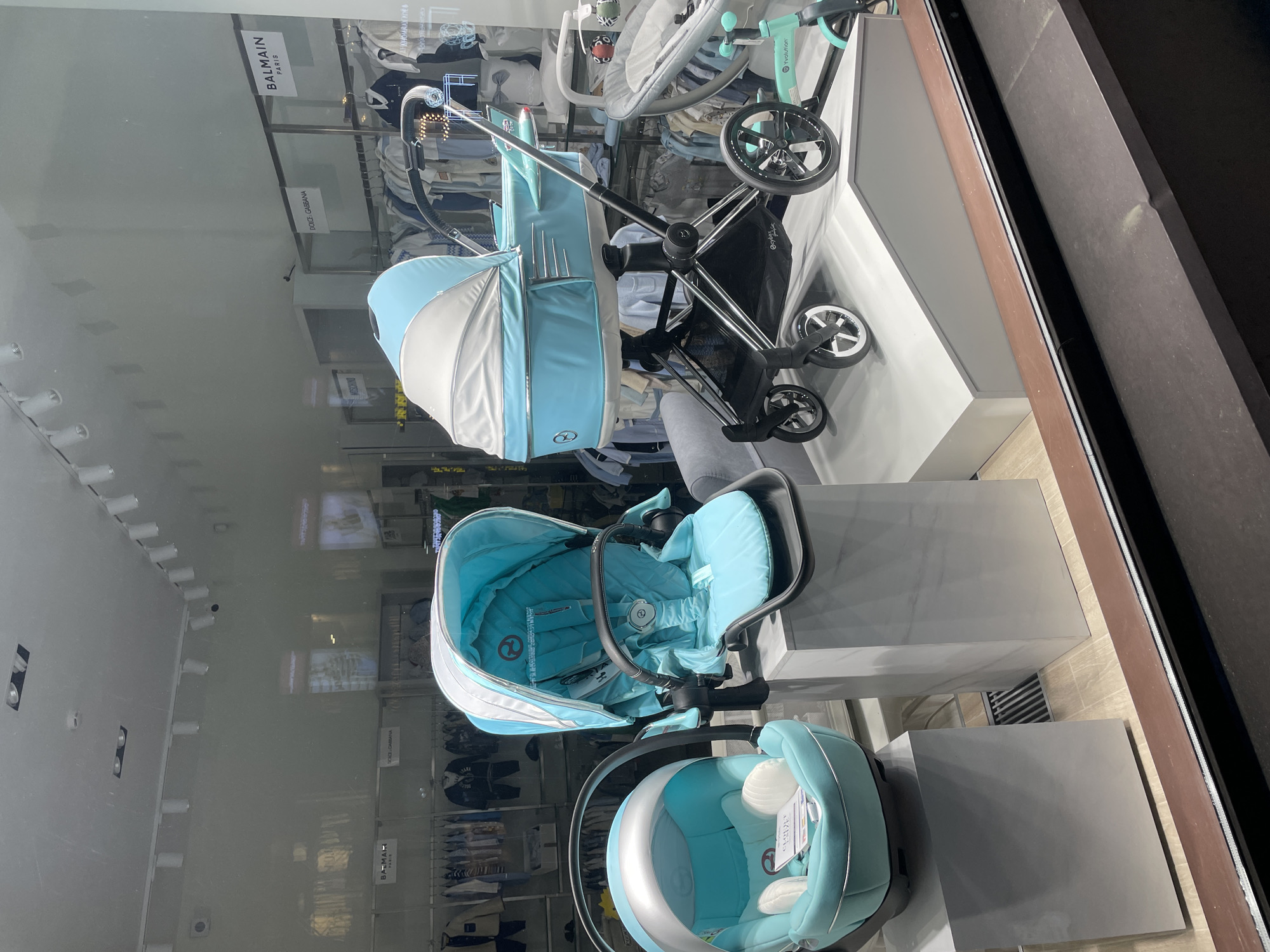 The image shows a retail display of baby strollers and car seats in a storefront window. The items are arranged on elevated platforms, presumably for better visibility. The strollers and car seats are light blue and white. The reflection in the window shows the interior of the store, including lighting fixtures and clothing racks. The brands "BALMAIN" and "DOLCE & GABBANA" are visible in the window reflection. The location of the image is unknown, so I cannot confirm it is in St. Petersburg, Russia.