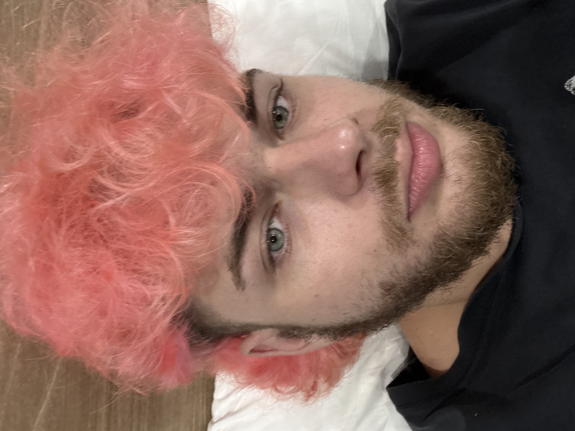 A close-up portrait captures a person with vibrant, voluminous pink hair, lying down and looking directly at the camera. The person has light skin, striking light green or hazel eyes, and a prominent brown beard and mustache. Their lips are a natural pink and slightly parted. They are wearing a dark, possibly black, crew-neck shirt. The setting appears to be indoors, with the person resting on a white pillow or sheet. A light brown wooden surface is visible to the left of their head. The image focuses on their facial features and upper body, with a slightly elevated camera angle. The person maintains a neutral to contemplative expression. No specific activity beyond posing for the photograph is evident. The time of day cannot be determined from the lighting. Although the context provided is Đà Nẵng, Vietnam, there are no specific visual cues within the image to confirm this location. No visible text is present.