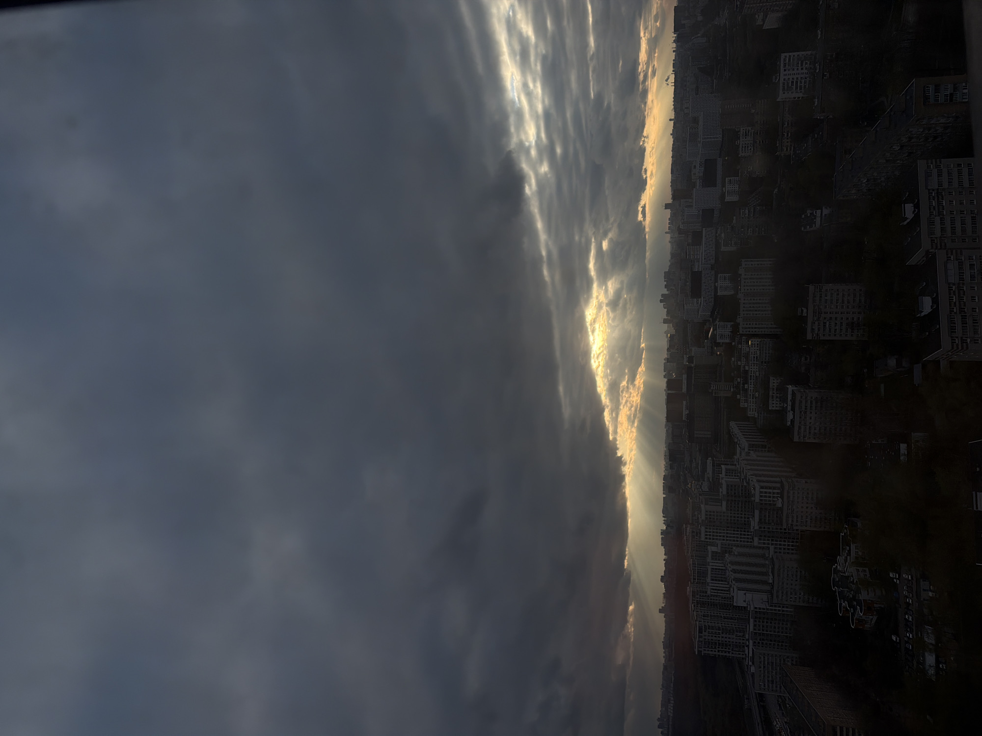 The image shows a view of Khimki, Russia. The sky is filled with dark, heavy clouds, partially obscuring the sun, which casts light rays down onto the cityscape. The city itself is composed of numerous buildings, primarily residential apartment blocks. The scene is viewed from an elevated position, giving an overview of the city extending towards the horizon.