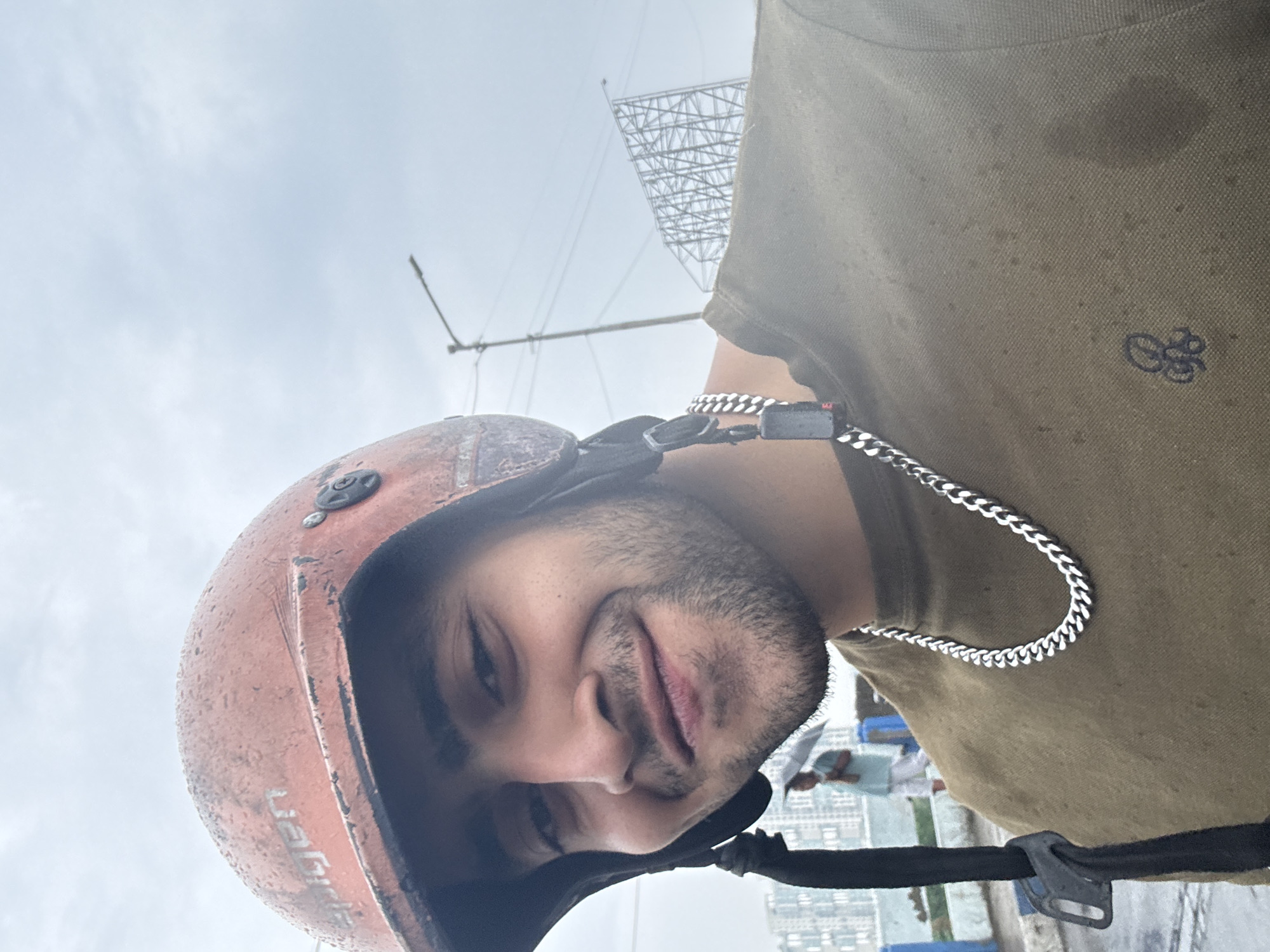 A man wearing a motorcycle helmet and an olive green polo shirt stands on a rooftop in Kolkata, India. He is facing the camera and appears to be smiling slightly. He wears a silver chain around his neck. The helmet is rusty orange in color and shows signs of wear. Raindrops are visible on the helmet and the shirt, suggesting wet weather. In the background, there is a structure, possibly scaffolding or a radio tower, and a cityscape with buildings. People are visible blurred in the background.