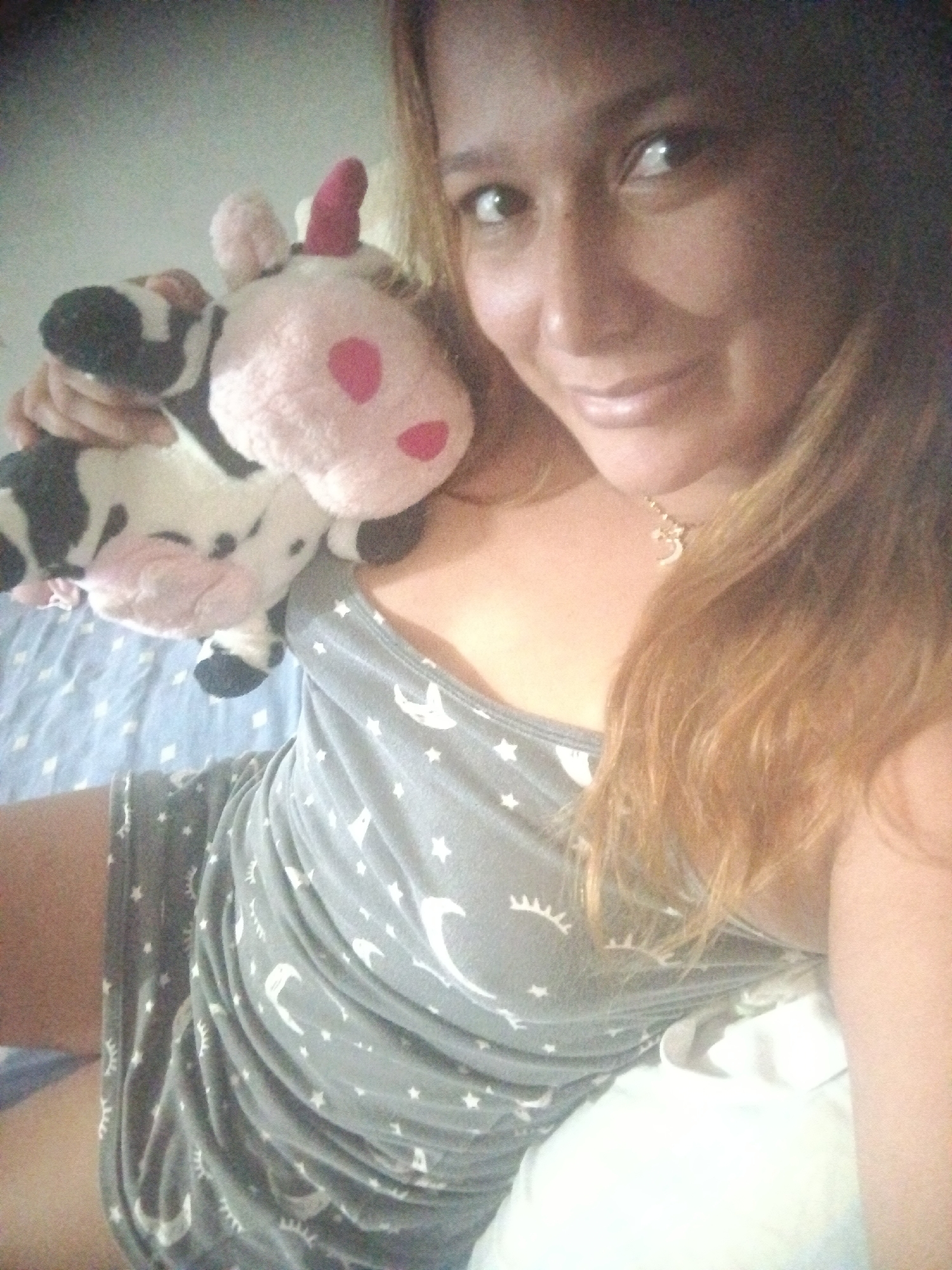 The image captures a smiling person, likely female, looking directly at the camera. She has long, reddish-brown hair that falls over her shoulders. She is holding a plush toy, which is a black and white cow with a pink face, pink spots, and a distinct pink horn, suggesting it might be a unicorn-cow hybrid.
The person is wearing a casual grey tank top or dress, patterned with white stars, crescent moons, and stylized eyelash symbols. A delicate golden necklace with a small charm is visible around her neck.
The background suggests an indoor setting, likely a bedroom, with a light blue patterned sheet or fabric visible on the left and a white pillow or bedding on the right. The person's posture and direct gaze indicate a relaxed pose, possibly a selfie. Her expression is calm and friendly, with a gentle smile. The image has a slightly grainy quality. There is no visible text within the frame. While the context states Caracas, Venezuela, no specific visual elements in the image directly confirm this geographical location.