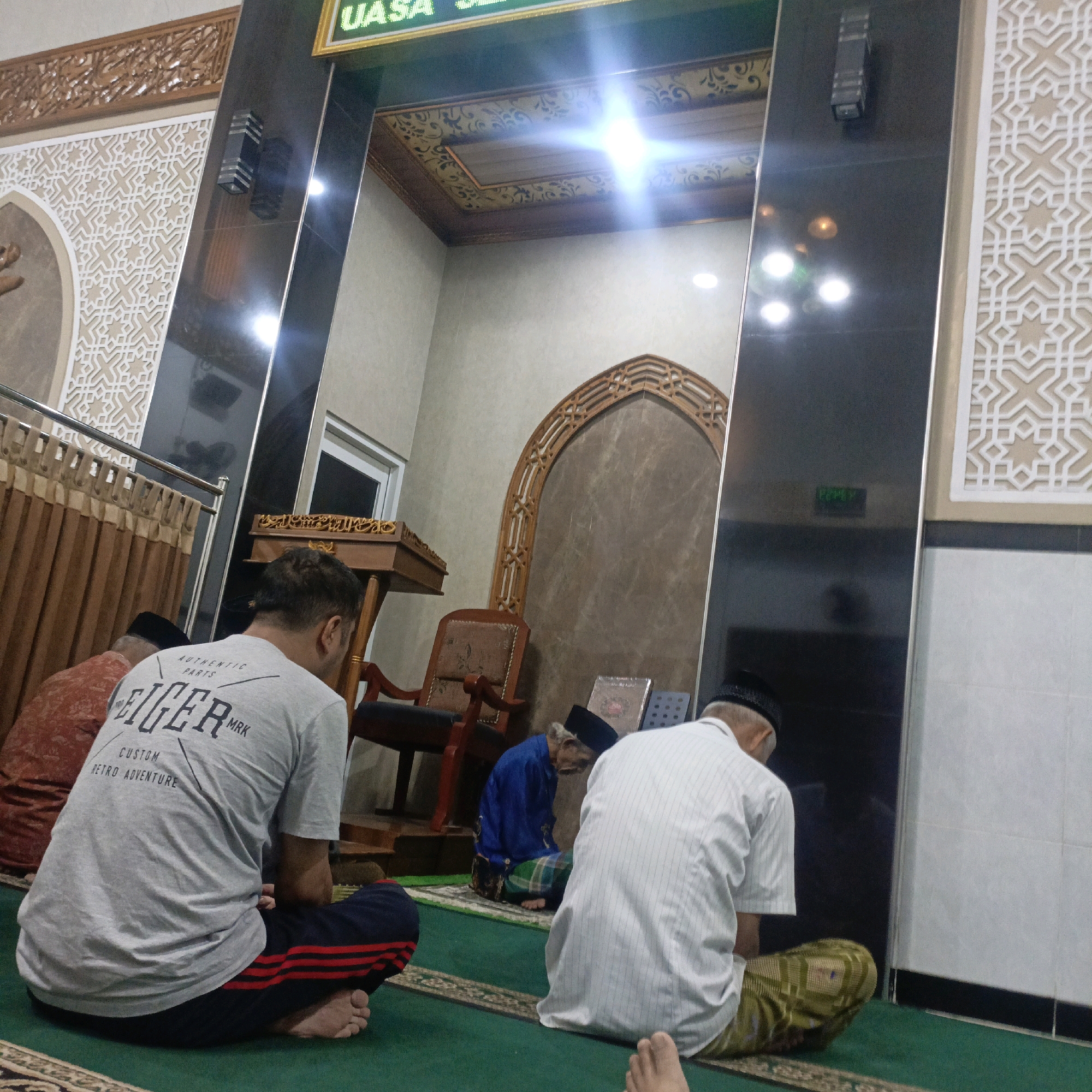 The image depicts an interior scene, likely within a mosque or prayer hall, in Salatiga, Indonesia. Several individuals are seen in prayer or seated on a green carpeted floor.
In the foreground, on the left, a person wearing a grey t-shirt with "EIGER MRK CUSTOM RETRO ADVENTURE" printed on the back is seated with legs crossed. They are wearing dark blue pants with red stripes. To their left, another person in a red patterned garment and a black skullcap is also seated.
In the mid-ground, a man in a blue shirt and a checkered sarong is seated, facing away from the camera and appearing to be in prayer. Next to him are two books. Behind this man, a wooden chair with dark upholstery is visible. To the left of the chair is a wooden podium with ornate carvings, possibly for religious texts.
On the right side of the image, a man in a white striped shirt and a patterned sarong is seated, facing away from the camera. His posture suggests he is also engaged in prayer.
The environment features ornate Islamic architectural elements, including patterned wall decorations, carved wooden accents around an arched doorway, and a decorated ceiling with intricate patterns. A large, dark, reflective surface, possibly a pillar or wall panel, is on the right side of the image, reflecting some of the lights and surroundings. Above the archway, a digital display with green text, possibly indicating prayer times or other information, is visible. The overall lighting suggests an indoor setting with artificial lights.
