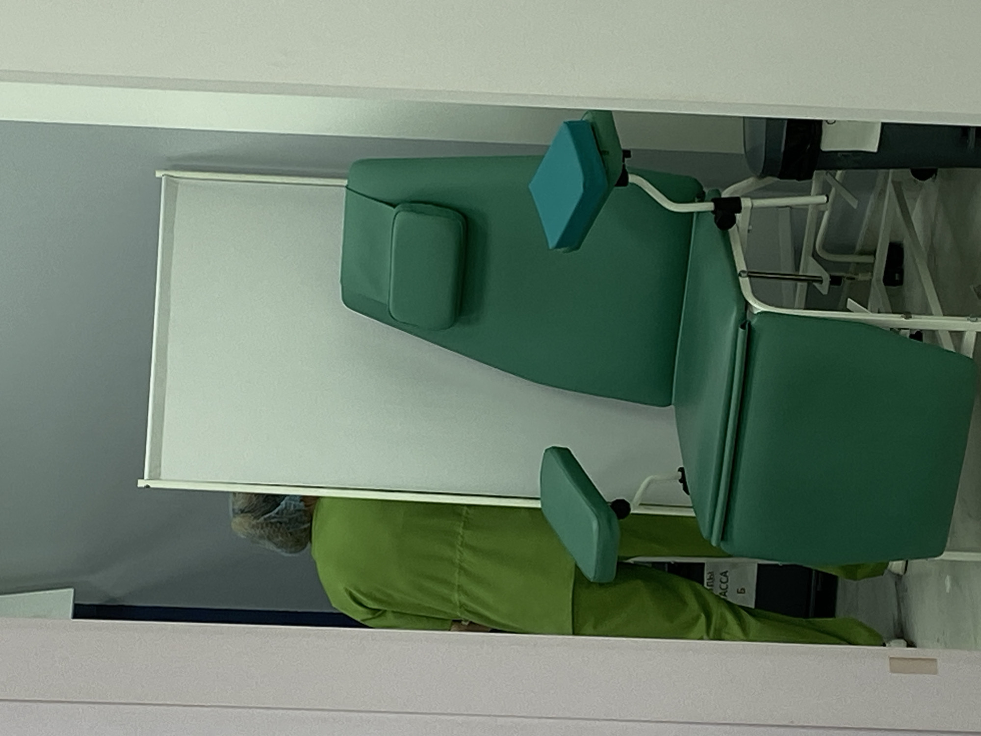 In Sokhumi, Abkhazia, a sterile medical room features a green reclining chair with adjustable armrests and a headrest, intended for patient procedures. Adjacent to the chair is a tall, white privacy screen. To the left, a person wearing green medical scrubs and a hairnet is partially visible, facing away from the camera. The flooring is a light grey. Partially obscured text on a black surface reads "ACCA". A dark gray trash receptacle with a black liner is situated to the right of the chair.