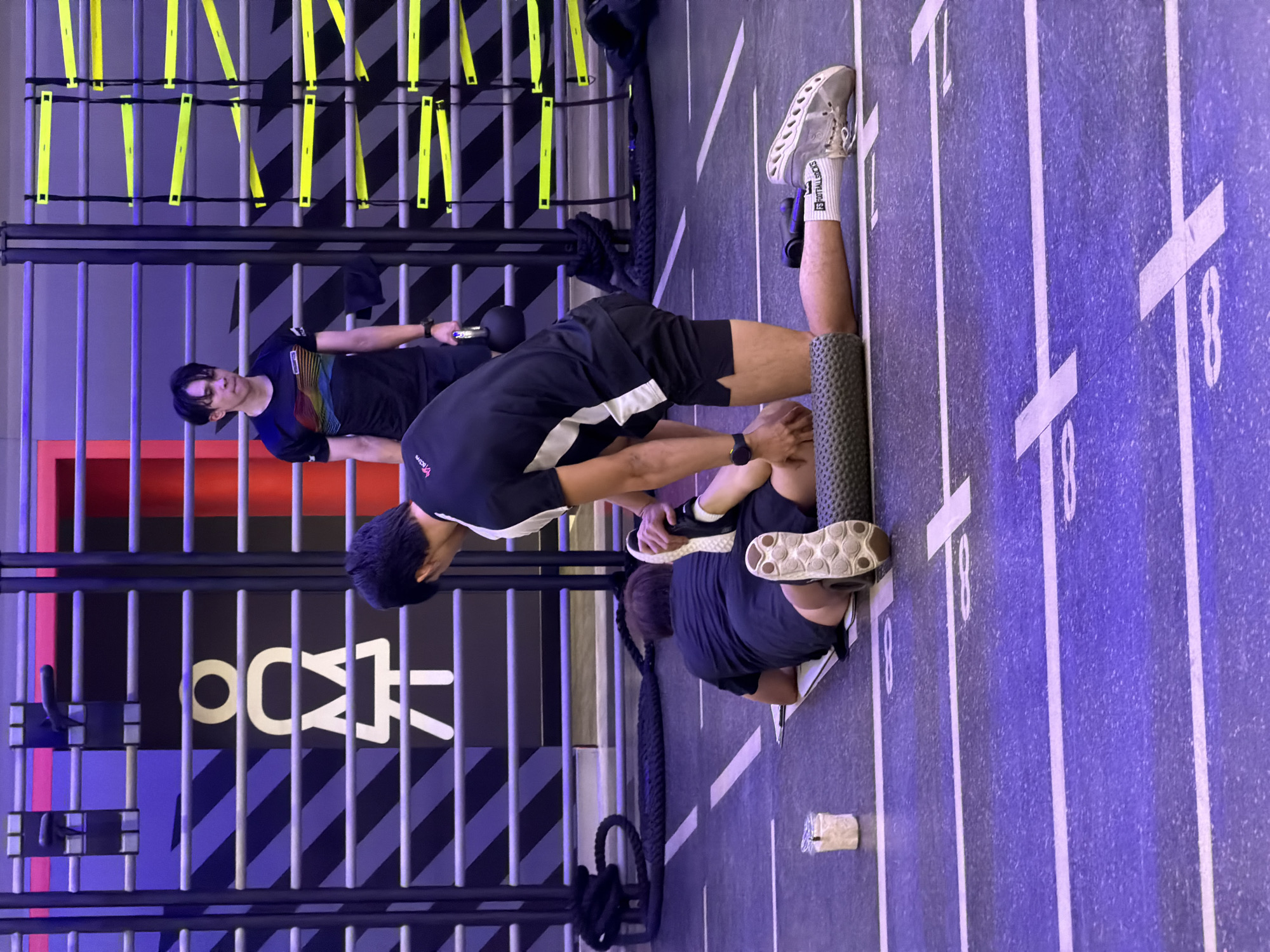 This image captures an active scene inside a modern fitness center or gym in Nong Pa Khrang, Thailand. The setting features a prominent blue-purple floor marked with white lines and numbers like "8" and "18," suggesting designated workout zones or lanes. The background wall is decorated with a striking black and dark grey striped pattern, behind which a metal cage-like structure with bright yellow suspension straps or resistance bands is visible.
Three men are engaged in various fitness activities. In the foreground, a man with dark hair, wearing a black and white athletic short-sleeved shirt and black shorts, is performing foam rolling on his left leg. He is using a grey textured foam roller and has white sneakers with "FOOTBALLSOCKS" clearly visible on his socks. To his left, slightly behind, another man in a dark blue short-sleeved shirt and black shorts is seated on the floor, appearing to stretch or rest, with his dark sneakers visible. A small white water bottle or cup is on the floor near him. Further in the background, a third man with dark hair is standing, wearing a multi-colored striped t-shirt and dark trousers, holding a dark kettlebell in his right hand, suggesting he is either about to begin or has just completed a strength exercise.
The lighting is bright and artificial, typical of an indoor gym environment. On the metallic structure in the mid-ground, a large stylized logo "O<Δ" is visible, possibly representing the gym's brand. The overall ambiance is one of focused exercise and training.
