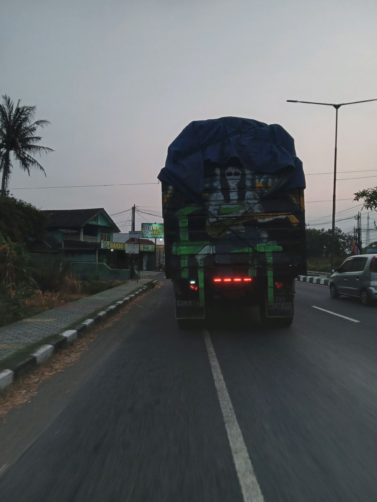 In Geneng, Indonesia, an image captures the rear view of a truck on a road during dusk. The truck is covered by a dark blue tarp, revealing a painted mural of figures wearing sunglasses and a guitar. Red brake lights are illuminated on the truck's rear. To the left of the truck, a sidewalk with a decorative border is visible, bordered by dry vegetation. Beyond the sidewalk, a light green building with a balcony stands, accompanied by signage for "BAKSO PAK NAN." Further down the road, a silver car is in the right lane. The sky is overcast, with a street lamp visible on the right side of the image.