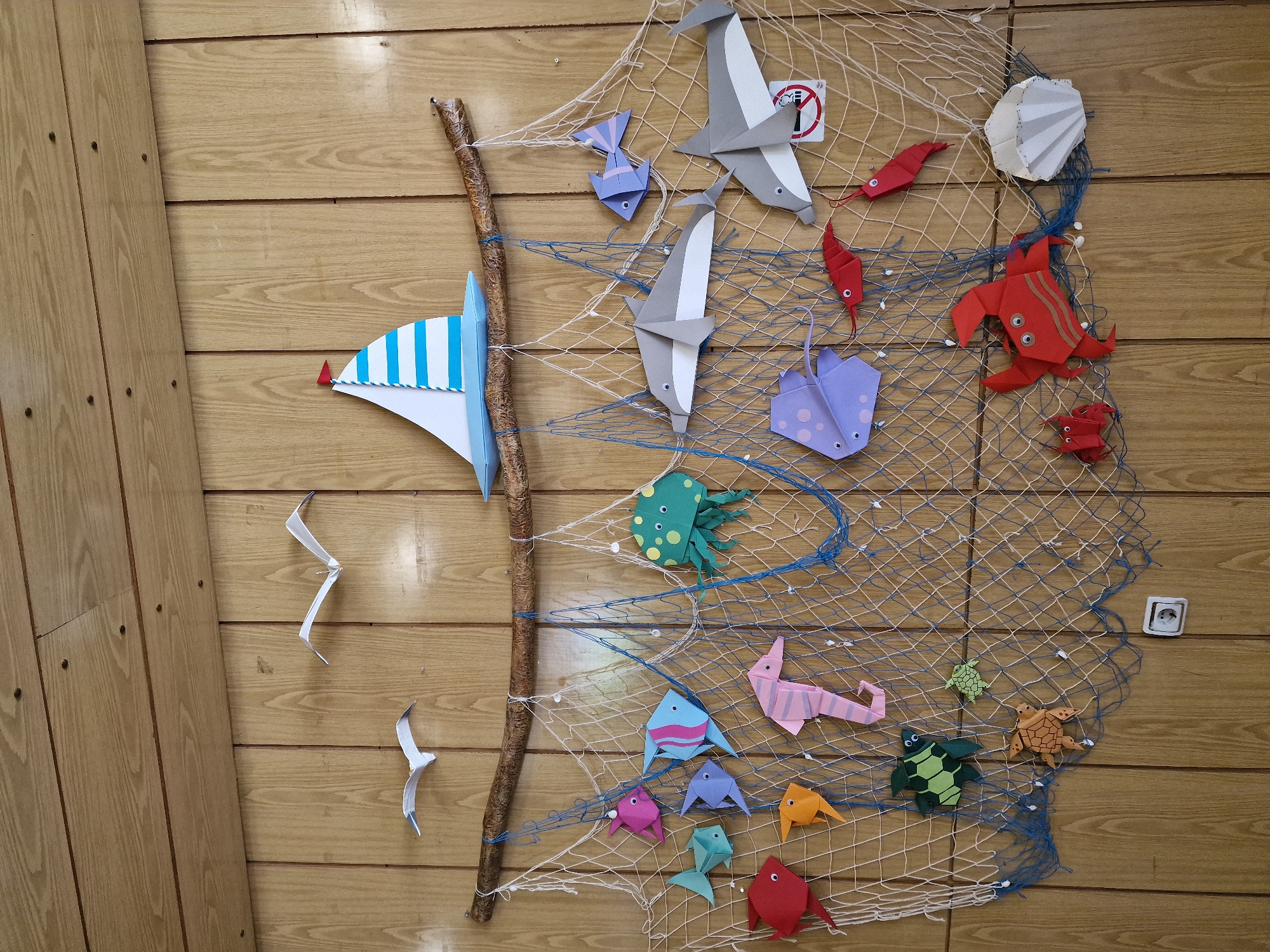 The image shows a wall decorated with origami sea creatures and a sailboat, suspended on a fishing net attached to a branch. The wall is made of light-colored wood paneling. At the top, two origami seagulls are attached to the wall. A blue and white striped sailboat with a red flag is positioned above the branch. The fishing net, made of white and blue netting, hangs down the wall and has various origami figures caught within its mesh. These figures include several fish in different colors (pink, blue, red, orange, green, and purple), a pink seahorse, a green octopus with yellow spots, two gray origami dolphins, a purple stingray, two red ribbon-like fish, a green turtle, a smaller green turtle, an orange crab, a small red crab, and a white origami seashell. The overall scene is a colorful and whimsical nautical-themed decoration. An electrical outlet is visible on the lower part of the wall.