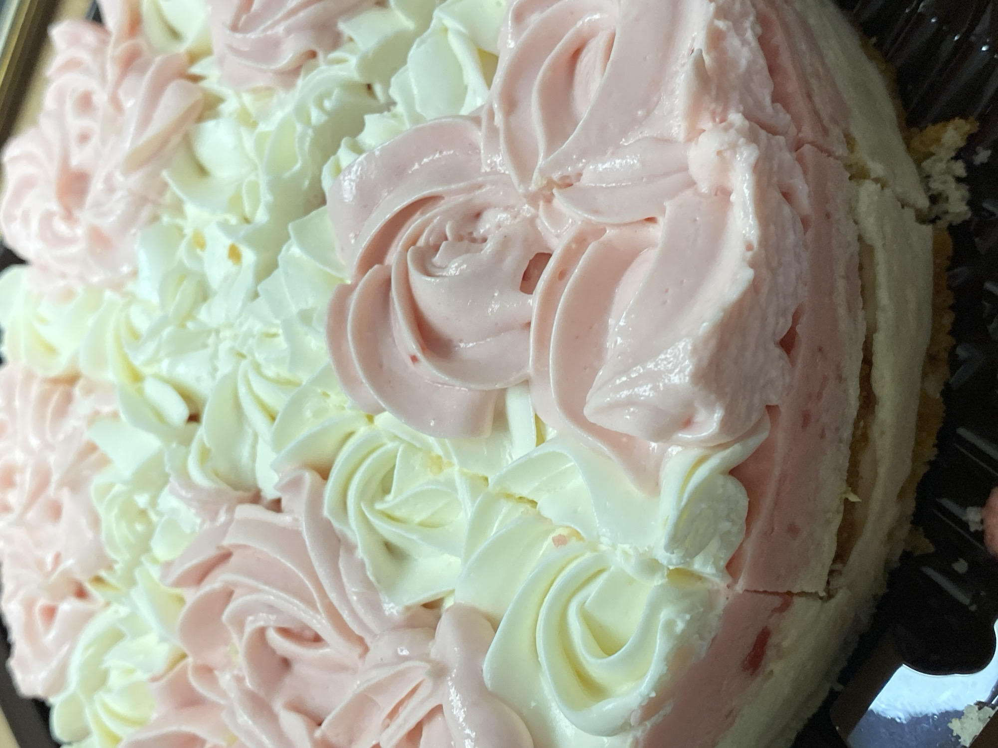 The image shows a close-up of a cake decorated with alternating rows of pink and white frosting rosettes. The cake appears to be circular and partially cut. The cake is in a black plastic container. There are no people visible in the image. There is no information to determine if this image was captured in Votkinsk, Russia.