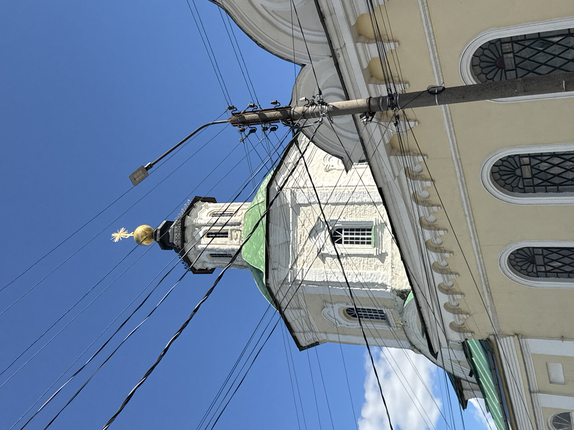 The image shows a view looking up at a church with a golden dome, situated against a bright blue sky. The church's architecture features a white facade with decorative elements and arched windows. A green roof is visible on one section of the building. A utility pole with numerous wires crisscrosses the frame, obscuring parts of the church and the sky. The wires are tangled and run diagonally across the image. The lighting suggests it is daytime, likely sunny, given the clear sky and shadows. The presence of the church with its dome and architectural style, combined with the provided location context of Yelets, Russia, indicates a religious building in that city. No people or specific events are depicted. The image is oriented sideways, with the church tower appearing to extend upwards from the bottom right of the frame.