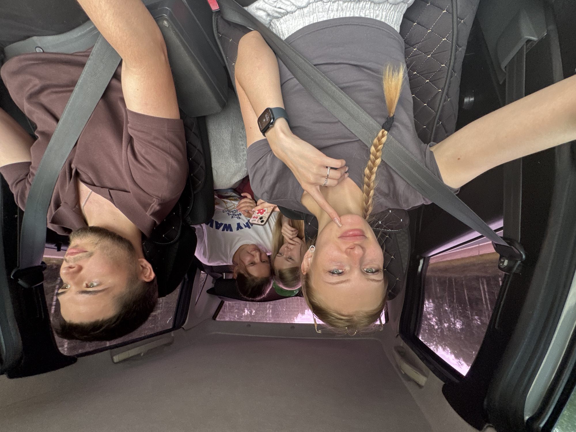 The image shows the interior of a car with four people. In the front passenger seat, a young woman with blonde braided hair is taking a selfie. She is wearing a grey t-shirt and a smartwatch. To her right, in the driver's seat, a young man with short dark hair is looking forward. In the back seat, two young women are visible. One is in the middle, looking at her phone, and the other is to her right, also looking at her phone. The car appears to be in motion, as evidenced by the trees and road visible through the windows, suggesting it's on a highway or a road outside of a city. The setting is a rural or semi-rural area with trees lining the road, consistent with the outskirts of Selishcha, Russia. The overall mood appears casual and familial.