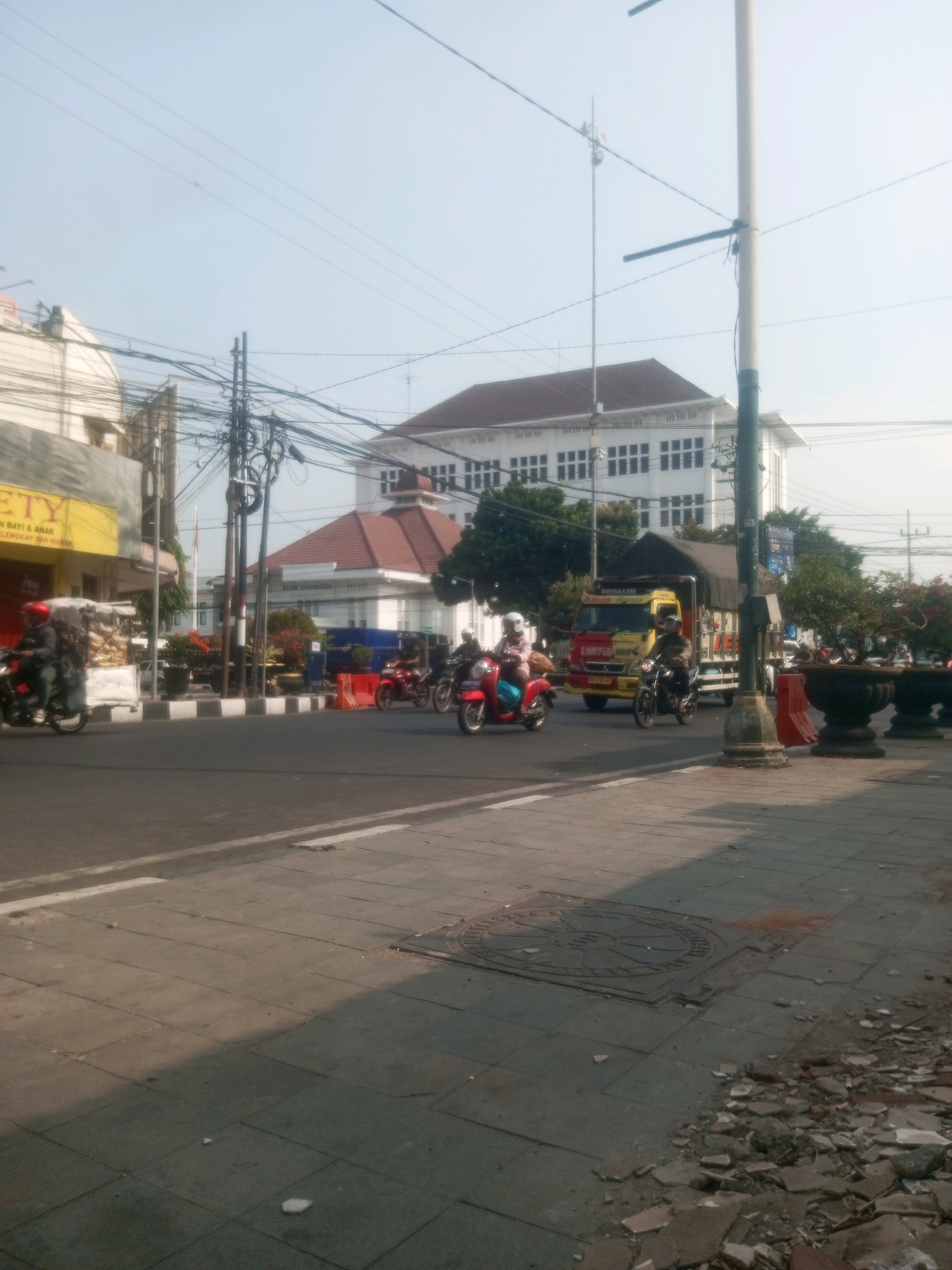 In Kediri City, Indonesia, a busy street scene unfolds under a clear sky. Several motorcycles are in motion, with riders wearing helmets. One motorcycle is carrying a load of goods. In the background, a yellow and red truck is visible, its bed covered with a dark tarp. Various buildings line the street, including a large white building with multiple windows and a building with a red-tiled roof. Power lines crisscross the sky, supported by utility poles. The sidewalk in the foreground is paved with square tiles, with some debris scattered on the ground. A manhole cover is visible on the sidewalk. The overall impression is one of daily life and activity in an urban environment.