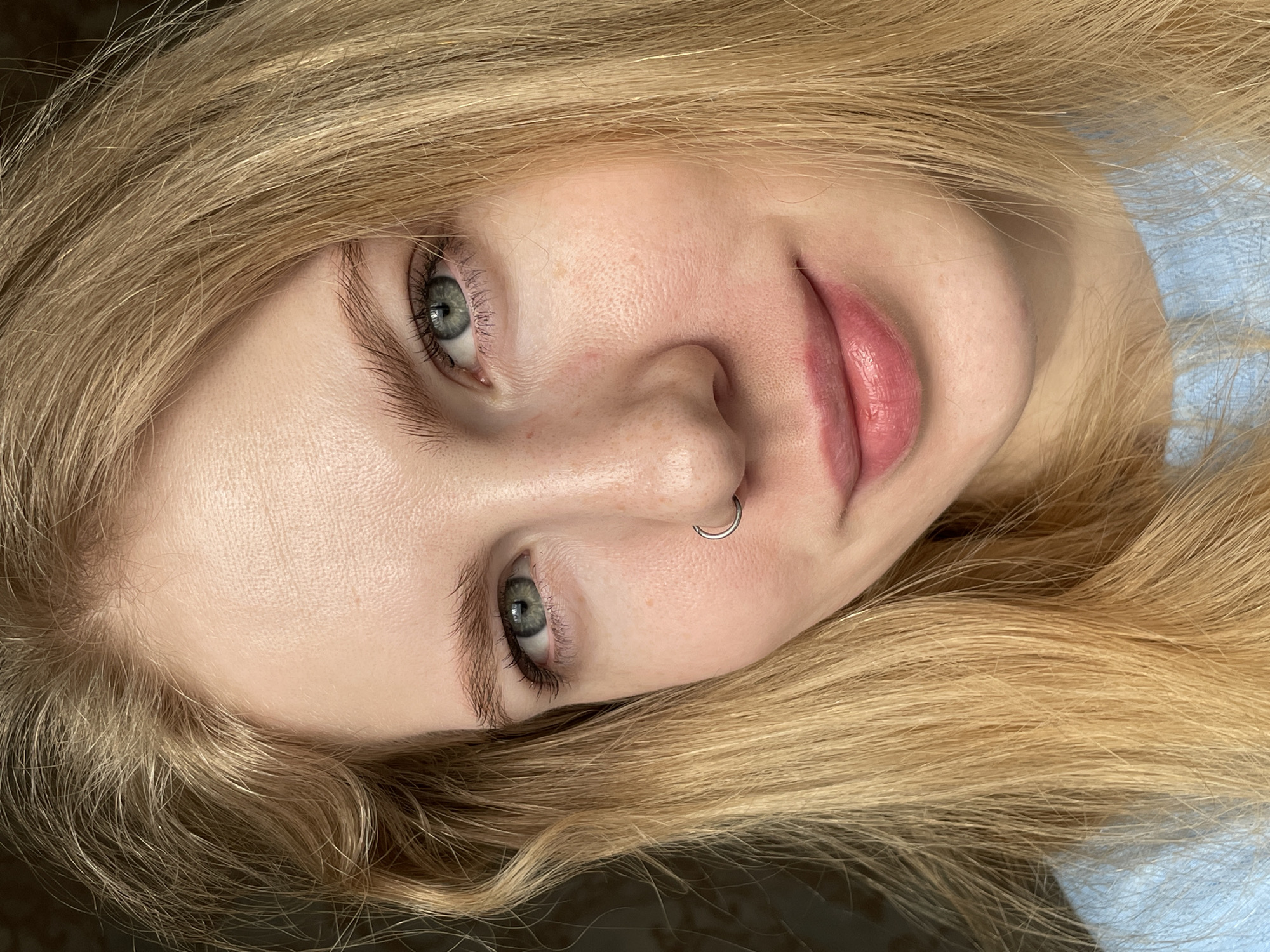 A close-up shot of a fair-skinned, blonde-haired woman with light green eyes. She has a silver nose ring in her left nostril. Her lips are a light pink color. The background appears to be a mix of shadows and light blue fabric. The scene does not provide enough information to determine if this photo was taken in the city of Izhevsk, Russia.