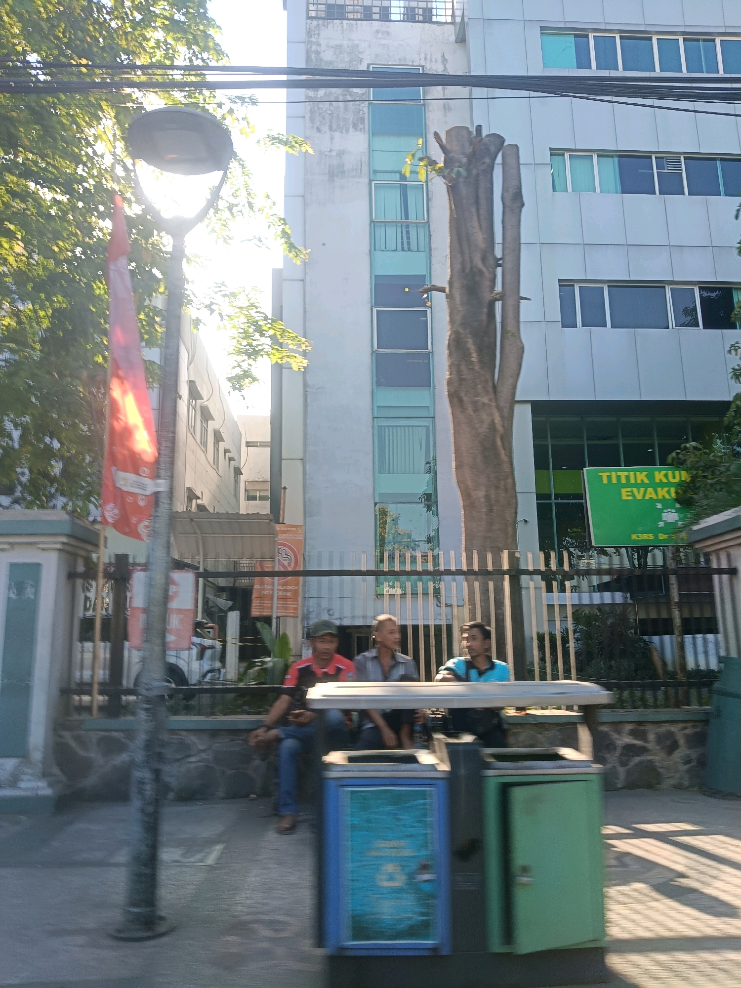 The image depicts a street scene in Surabaya, Indonesia, with a modern building as the backdrop. The building is a light gray, multi-story structure with numerous windows, some of which are tinted green and blue. In front of the building, a large, bare tree trunk stands prominently. A black metal fence separates the sidewalk from the building's grounds.
On the sidewalk, three men are seated on a bench. The man on the left wears a red and black shirt and blue jeans. The man in the middle wears a gray shirt and dark pants. The man on the right wears a blue shirt and dark pants. They appear to be resting or waiting. Two trash receptacles are visible on the sidewalk in front of the bench; one is blue and white with graphics, and the other is green with an open door.
To the left of the men and bench, a tall lamppost with a large lamp fixture stands, partially obscured by a red flag on a pole. Sunlight streams through the leaves of a tree on the left side of the frame, creating bright highlights. A green sign with white text that reads "TITIK KUMPUL EVAKUASI" (Evacuation Assembly Point) is attached to the fence in front of the building. The ground in the foreground is paved, and shadows from overhead wires and the tree stretch across it.