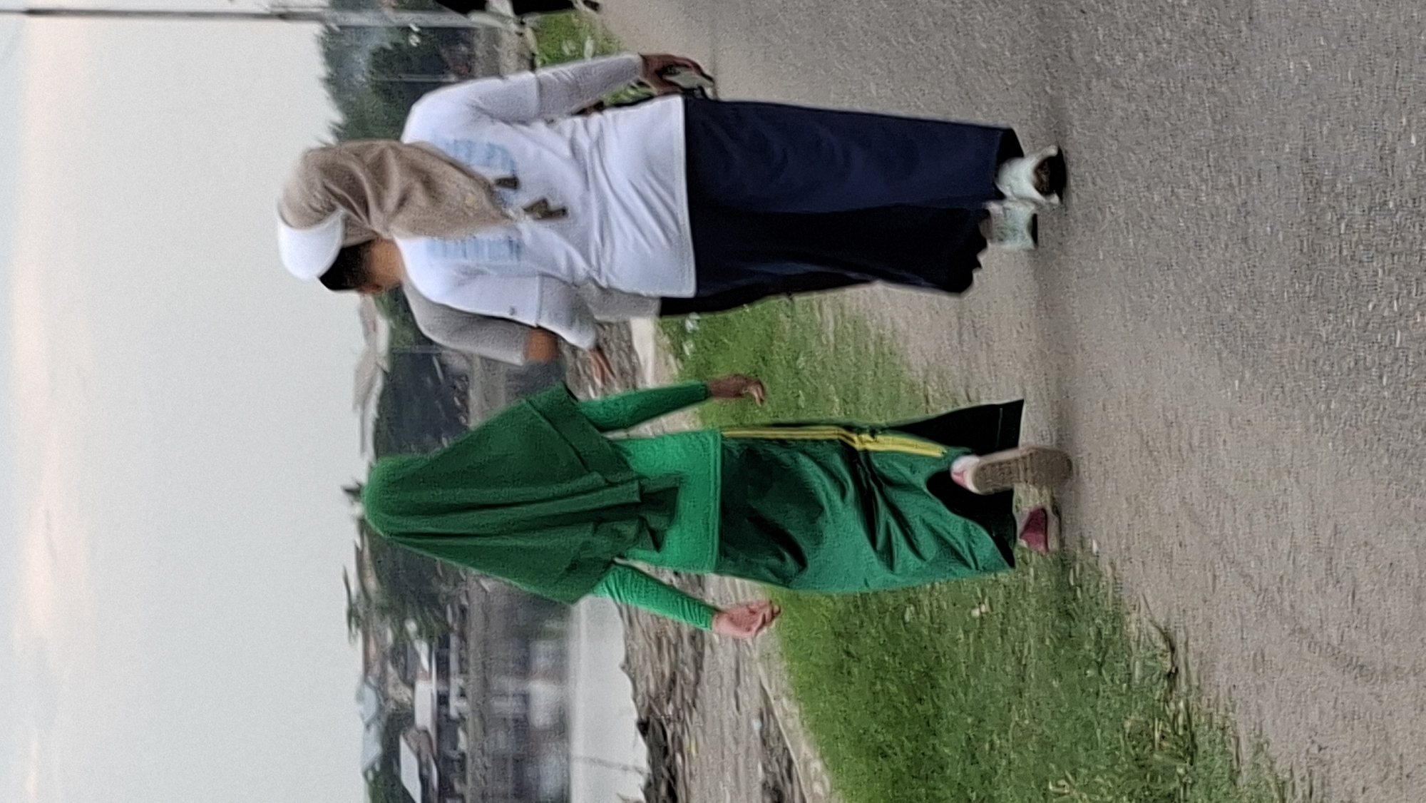 The image shows three individuals walking on a paved path in Lhokseumawe, Indonesia. The person in the foreground on the left is wearing a green hijab and a long green skirt with yellow stripes. They are walking away from the camera. To their right and slightly behind is another person wearing a grey t-shirt. Further to the right and behind is a third individual wearing a white long-sleeved shirt with a light brown hijab. This person is also walking away from the camera. The path is adjacent to a grassy area on the left, which slopes down towards a body of water with a line of buildings and trees in the background. The sky is overcast.