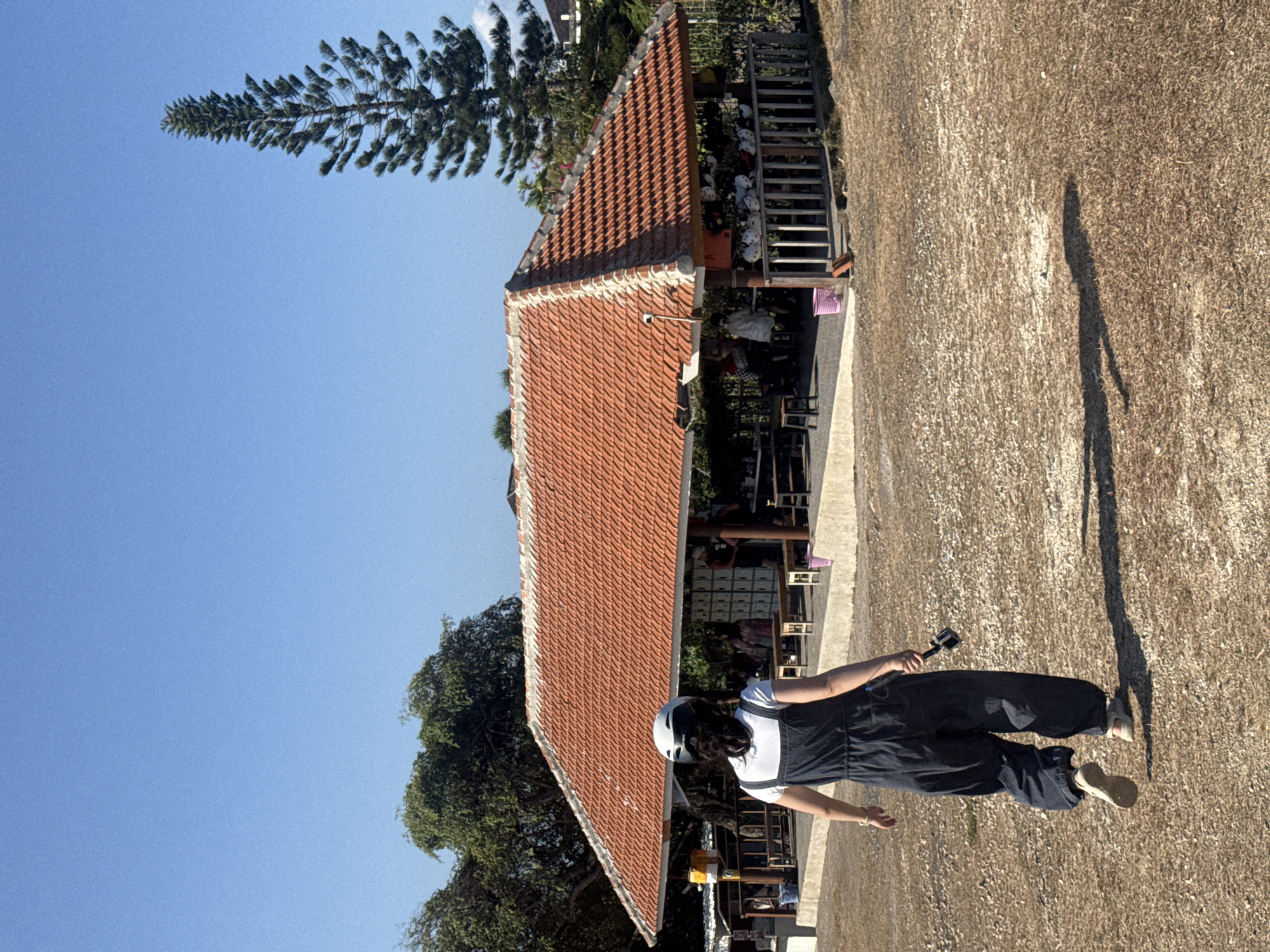 A woman wearing a white t-shirt and dark blue overalls, with a white and black helmet on her head, is walking away from the camera on a dry, grassy area. She is holding a black object in her right hand. In the background, there is an open-air pavilion with a red tile roof and wooden pillars. Several people are visible under the pavilion, and there are tables and benches. A tall, slender pine tree stands to the right of the pavilion. The sky is clear and blue, and the scene appears to be outdoors in a sunny environment, likely in Kuta Selatan, Indonesia, given the architectural style and vegetation.