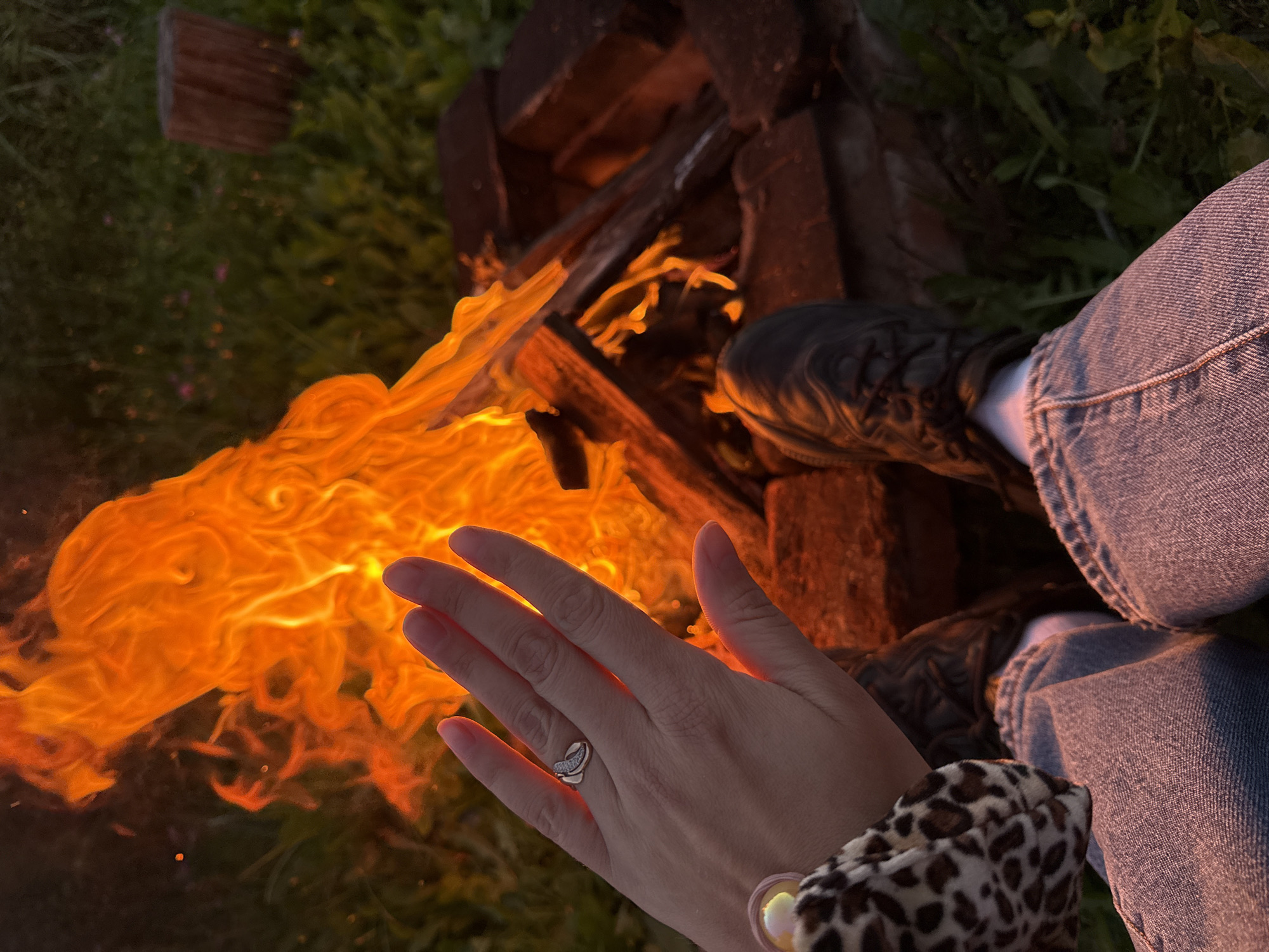 An outdoor scene features a person sitting by a campfire. The person's hands are visible in the foreground, with a ring on one finger, and they are wearing blue jeans and dark shoes. A cheetah-print accessory is also visible. The campfire is in the center of the frame, with bright orange flames and logs burning. The surrounding environment includes green grass and a wooden post in the background. The scene appears to be set in the evening or at dusk, with the fire providing the primary light source. The location is identified as Imelevo, Russia.