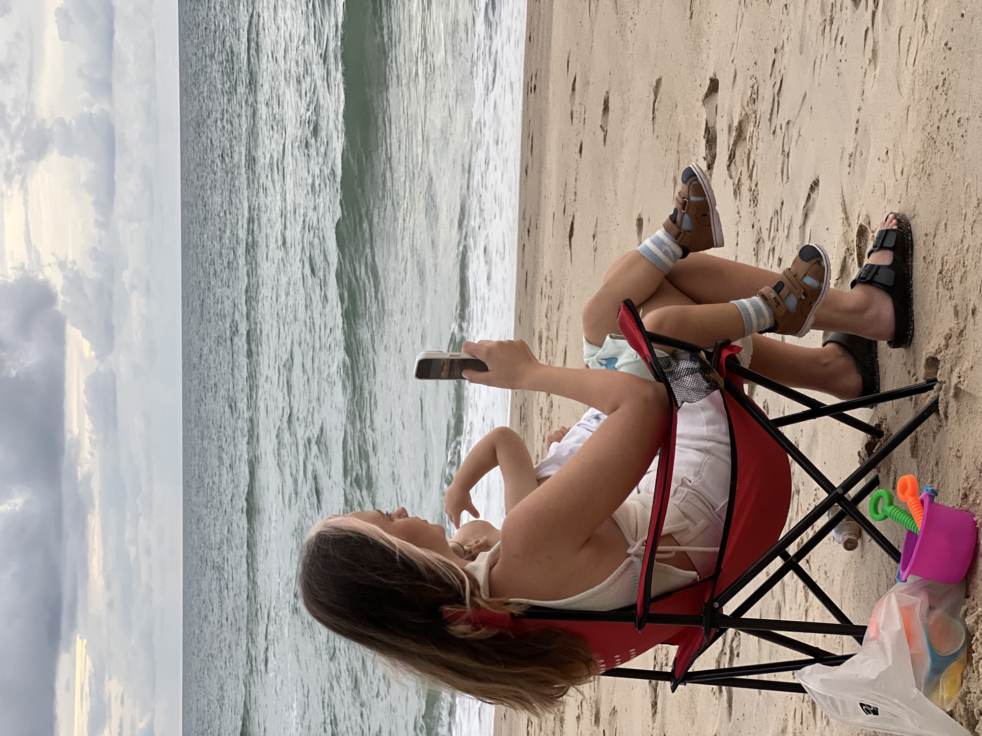 A woman is sitting in a red beach chair on a sandy beach, holding a mobile phone and looking at it. She is holding a baby in her lap. The woman has long, dark hair with some blonde streaks. She is wearing a white top and white shorts. The baby is wearing a light blue top. The woman is wearing black sandals and the baby is wearing brown sandals with blue and white striped socks. In front of the chair, there is a pink bucket with green and orange sand toys, and a white plastic bag with other beach toys. The ocean is visible in the background with waves. The sky is cloudy.