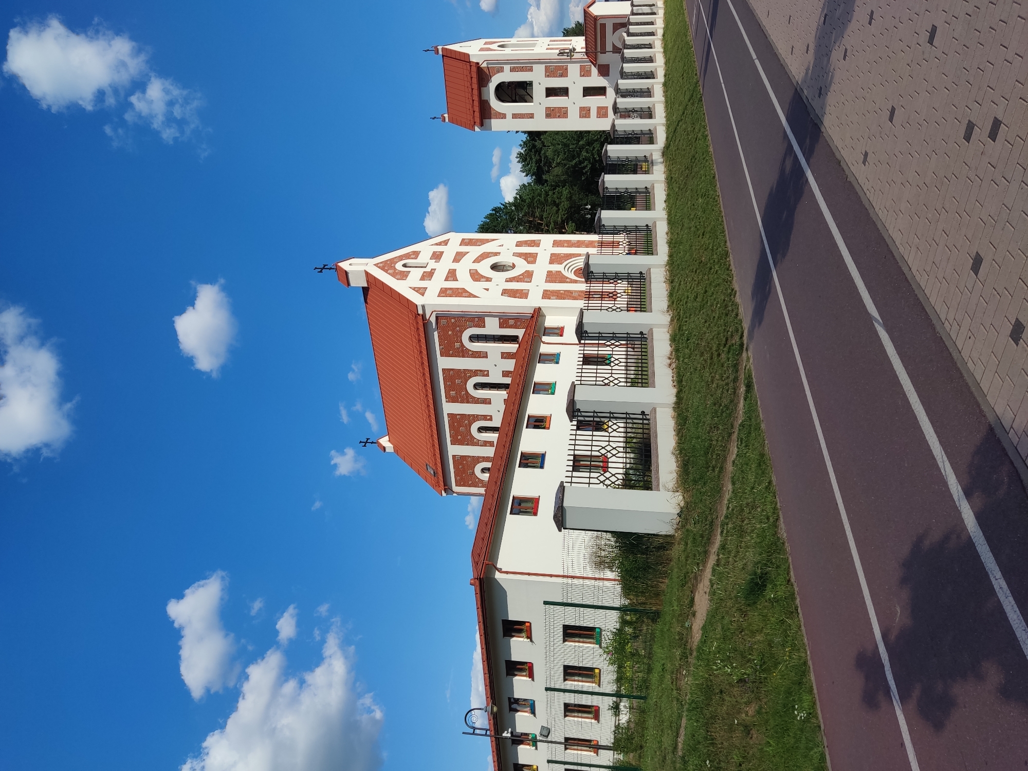 This is a wide shot of a religious complex with a church and a bell tower, located in the vicinity of the Бараўлянскі сельскі Савет, Belarus. The buildings are characterized by white walls and red tiled roofs, with distinctive red and white decorative brickwork on the church and bell tower. A low white fence with black metal railing separates the complex from a paved pathway and a road. To the left of the church, there is a white building with multiple windows, each featuring colorful frames. The sky is bright blue with scattered white clouds. The foreground shows a dark red asphalt path with two white lines, likely a bike path or pedestrian walkway, and a textured brick sidewalk. The lighting suggests it is daytime, with shadows cast on the path. No people are visible in the image.