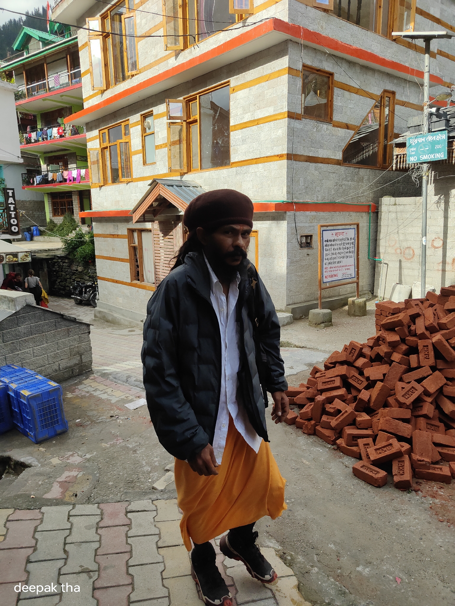 A man wearing a brown turban, a dark jacket, a white shirt, and an orange dhoti is walking on a paved walkway in Vashisht, India. He is looking towards the camera. To his right is a large pile of red bricks. Behind him is a multi-story building with a grey stone facade and orange trim. A sign is posted on the building that reads "No Smoking" in English and Hindi, along with a fine of 200 rupees. To the left of the man are blue plastic crates. Further in the background, more buildings with laundry hanging on balconies are visible, as well as trees and a mountain. A motorcycle is parked in an alleyway to the left. The sky is overcast.