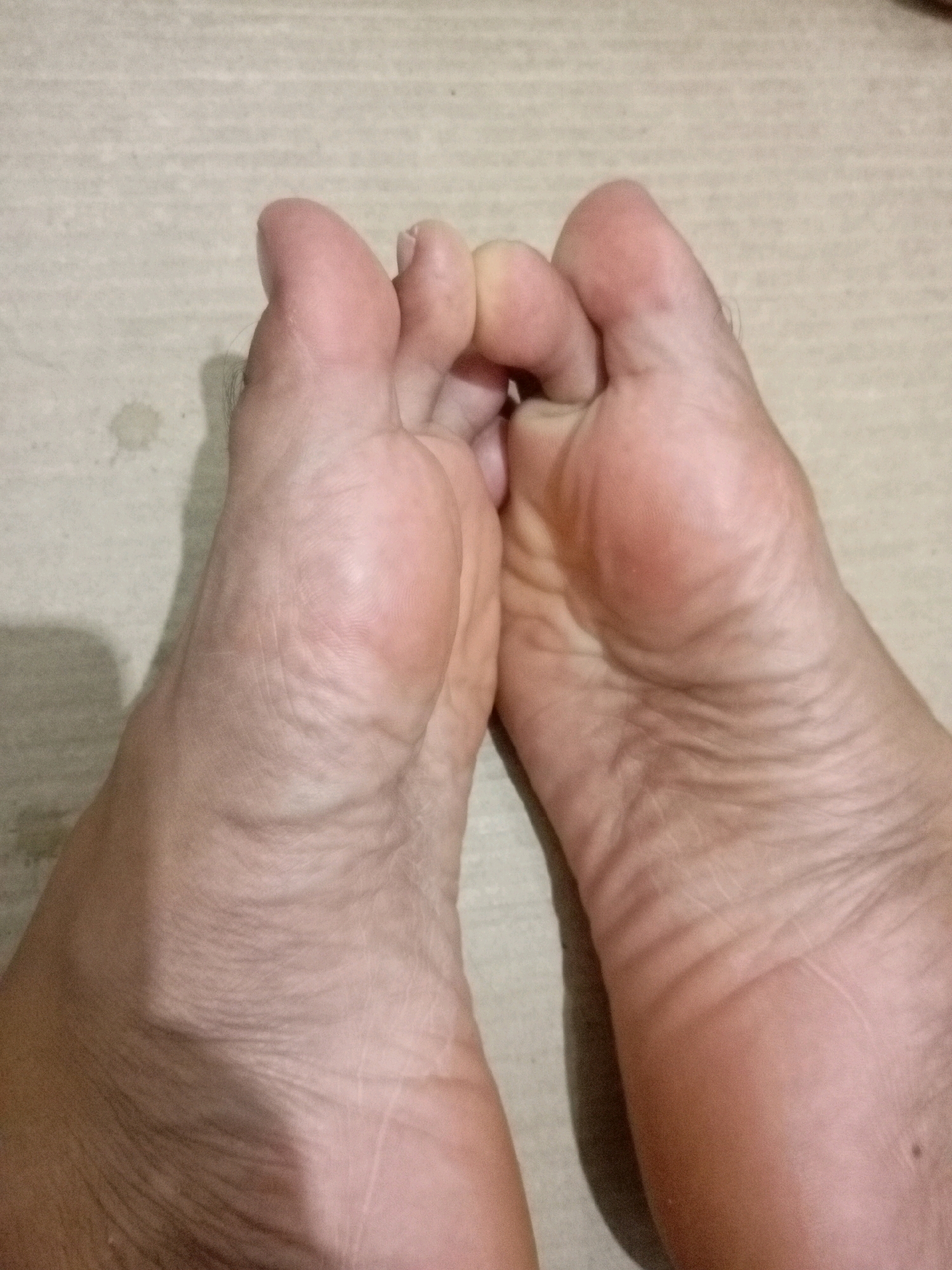 A close-up, eye-level, overhead shot shows two bare human feet positioned close together on a light-colored, textured surface. The feet are facing upwards and angled towards the viewer, with the toes of both feet interlocked. The skin on the feet appears dry and wrinkled, with visible lines and creases. The lighting creates a shadow on the left foot. The surface resembles a tiled floor or a similar material common in indoor environments. The location is identified as Batam, Indonesia.
