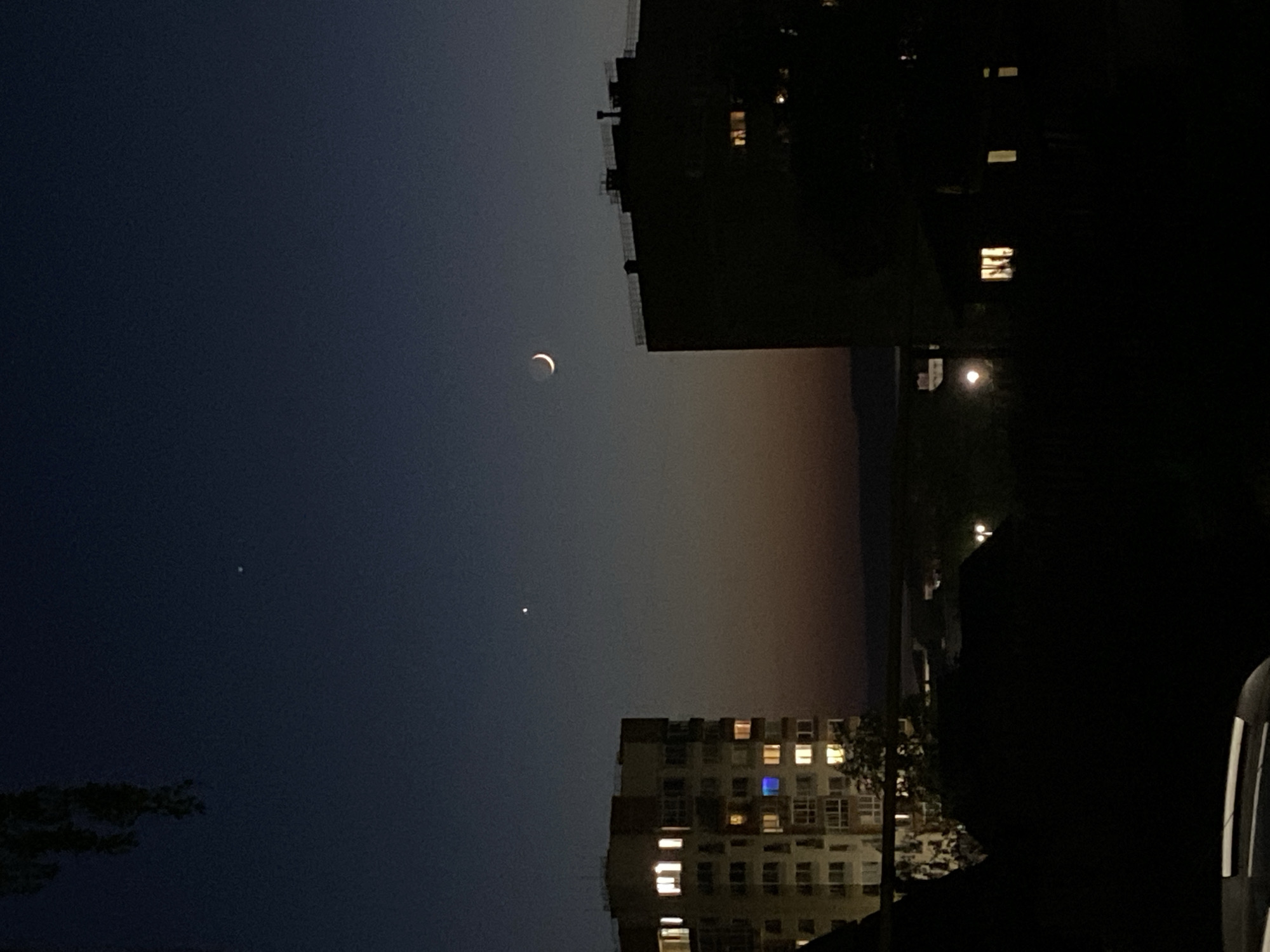 The image depicts an evening scene in Votkinsk, Russia. A thin crescent moon is visible in the upper-left portion of the sky, with a single star or planet (possibly Venus) appearing below and to the left of the moon. Two apartment buildings are visible: one in the lower-center, with illuminated windows, and another in the upper-right, appearing mostly silhouetted. The sky has a gradient of color, transitioning from a dark blue near the top to a lighter, hazy orange closer to the horizon. The foreground is largely dark, with shapes of foliage and what may be a parked car visible. A streetlamp is visible on the far right, near the horizon line.