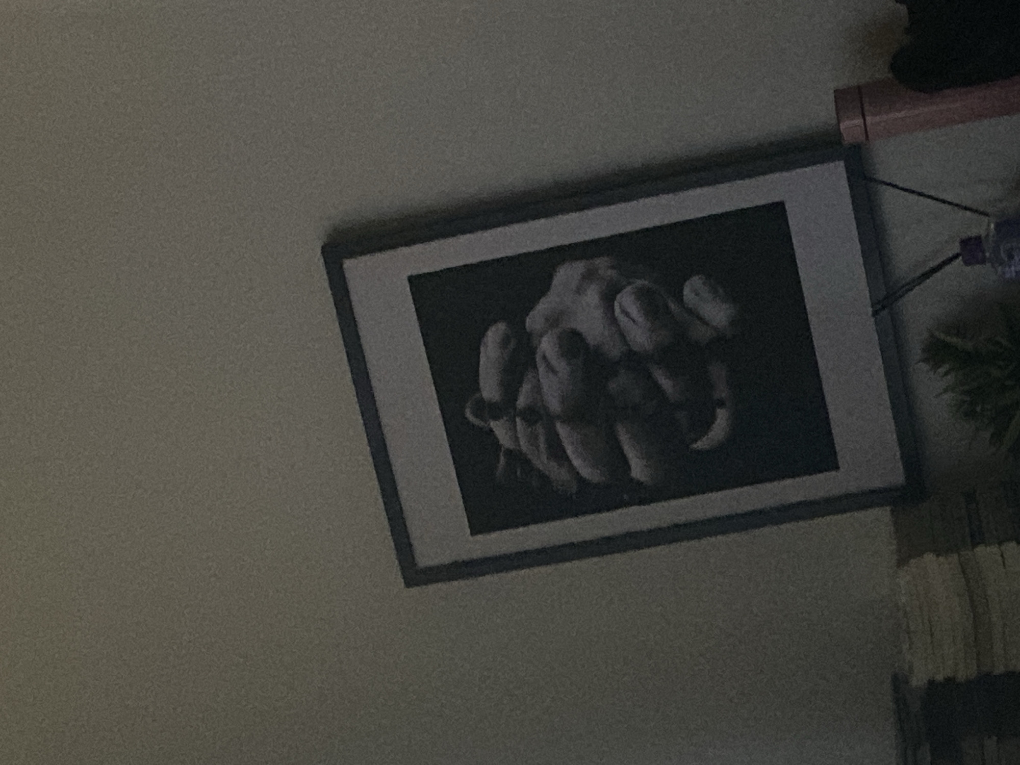 The image presents an interior scene, likely a room in Jos, Nigeria, captured from a low, upward-angled perspective. Dominating the view is a framed picture hanging on a light-colored, textured wall. The picture itself is a black-and-white photograph, depicting an abstract arrangement of what appears to be intertwined human hands or fingers. These hands are closely clustered, creating a complex, organic shape against a dark background, with lighting highlighting the contours and textures of the digits. The frame surrounding this artwork is dark.
To the right of the framed picture, several other objects are partially visible. A light-brown or wooden shelf-like structure is present along the top right edge. Below this, thin wires or strings descend, connecting to a small, purple object and further down to a dark green, textured item, possibly a hanging plant or a decorative tassel. In the bottom right corner, a segment of a striped or ridged fabric, potentially a curtain or upholstery, is visible.
The scene is static, with no discernible activity or event taking place, and no people are present. The lighting is dim, contributing to a noticeable graininess throughout the photograph, suggesting low-light conditions, possibly evening or a poorly lit interior during the day. No visible text is present within the image.