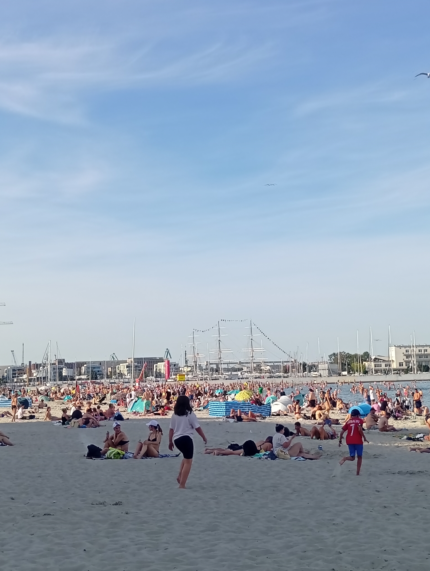The image depicts a crowded beach scene in Gdynia, Poland. A large number of people are sunbathing, sitting, and walking on the sandy shore. Some individuals are in the water, suggesting it is a popular swimming spot. In the background, a harbor with numerous boats, including large sailing vessels and smaller yachts, is visible. Cranes and buildings line the waterfront, indicating an urban setting adjacent to the beach. The sky is clear with a few scattered clouds. The overall atmosphere appears to be one of leisure and recreation on a sunny day.