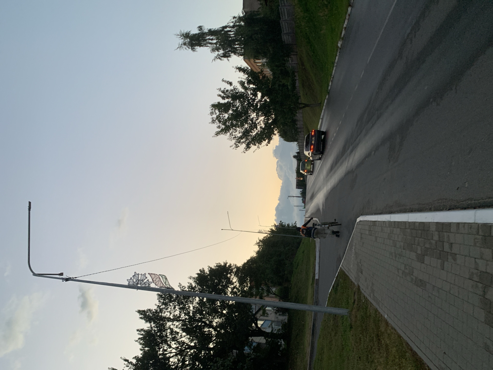 The image depicts a scene at dusk on an asphalt road in Kapyĺ, Belarus. A person is walking with a bicycle on a paved sidewalk to the left of the road. Further down the road, two cars are traveling away from the viewer. The sky is a pale blue with a gradient transitioning to a pale yellow near the horizon, indicating the time of day. Trees line the right side of the road, and a few buildings are visible in the background on the left. A street lamp with decorative elements is prominent on the left side of the image. The road appears to be slightly wet, with tire tracks visible. The environment suggests a suburban or semi-rural area on the outskirts of the city.
