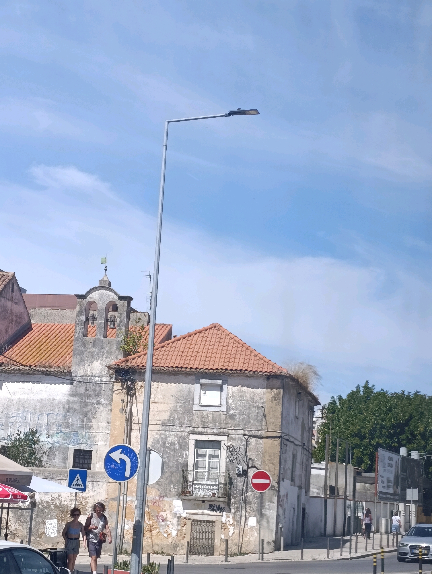 This is a medium shot of a street scene in Setúbal, Portugal. The image captures a view of old buildings with weathered facades and tiled roofs. A tall, modern lamppost stands prominently in the foreground, extending upwards towards a clear blue sky with wispy clouds.
To the left, a multi-arched bell tower, possibly part of a church, is visible, with bells hanging inside. A small green weather vane tops the tower. Adjacent to it is another building with a red-tiled roof. Graffiti is visible on the walls of some buildings.
Several street signs are present, including a blue sign with a white arrow indicating a left turn, a blue pedestrian crossing sign, and a red circular sign with a horizontal white bar, signifying "no entry" or "do not enter."
Two people are walking on the sidewalk in the mid-ground, seemingly engaged in conversation. One is wearing a light-colored top and shorts, while the other is wearing a dark top and shorts. They appear to be casually strolling.
In the background, a dense green tree line is visible, and behind that, some modern structures and billboards. A silver car is parked on the street in the lower right of the frame, and another car is partially visible on the left. The street itself is paved, with a sidewalk running alongside the buildings. The overall impression is of a sunlit day in an older part of a Portuguese city.