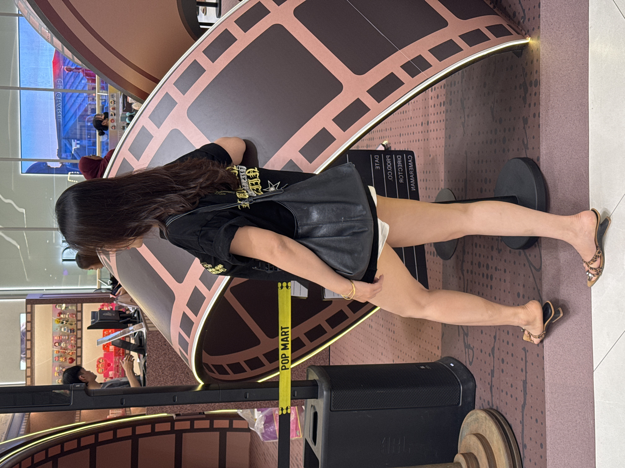 The image shows a woman standing next to a large, curved structure resembling film reel. She is wearing a black t-shirt, white shorts, and leopard-print slide sandals. She has a black shoulder bag and a gold bracelet on her left wrist. Behind her, there is a yellow tape barrier with "POP MART" written on it, and a black speaker on a stand. Further in the background, a man is seen working behind a counter in a store that displays various collectible figures. The overall environment appears to be an indoor commercial space, possibly a shopping mall, with patterned flooring and bright lighting. The sign "POP MART" suggests the location is a retail store. This scene is likely at a Pop Mart store in a shopping mall in Hanoi, Vietnam.