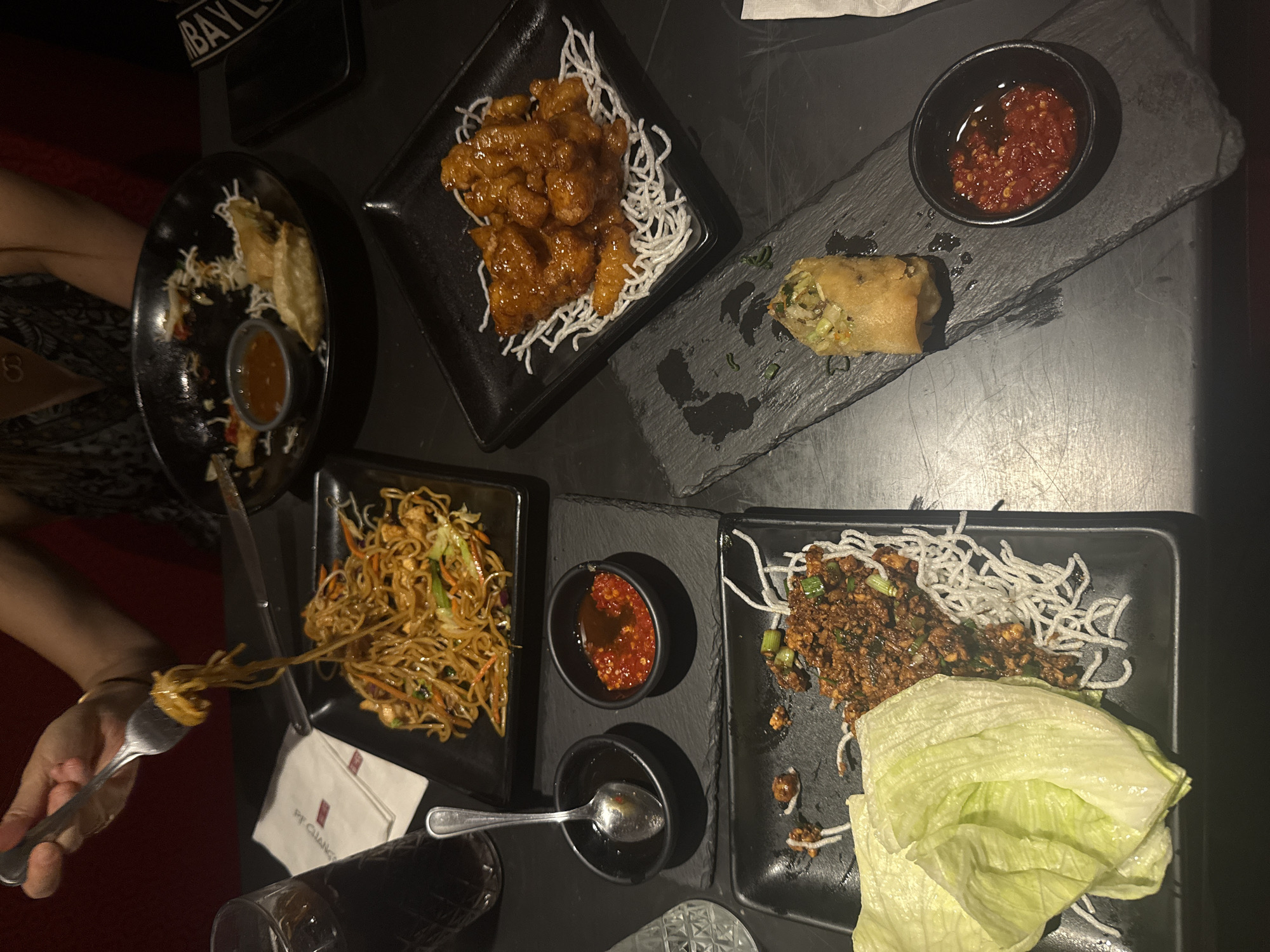 The image shows a table laden with dishes of food. Clockwise from the top left, a plate of dumplings with a dipping sauce, a plate of orange chicken with rice noodles, a slate board holding a spring roll and a small bowl of chili oil, and a plate of lettuce wraps with rice noodles. There is also a plate of chow mein noodles. A fork is piercing some of the noodles. A glass and a napkin with the PF Chang's logo is also on the table. Based on the logo, the food is being eaten at PF Chang's, although the specific location in Mexico City, Mexico cannot be determined from the image alone. A person's arms are visible on either side of the image.