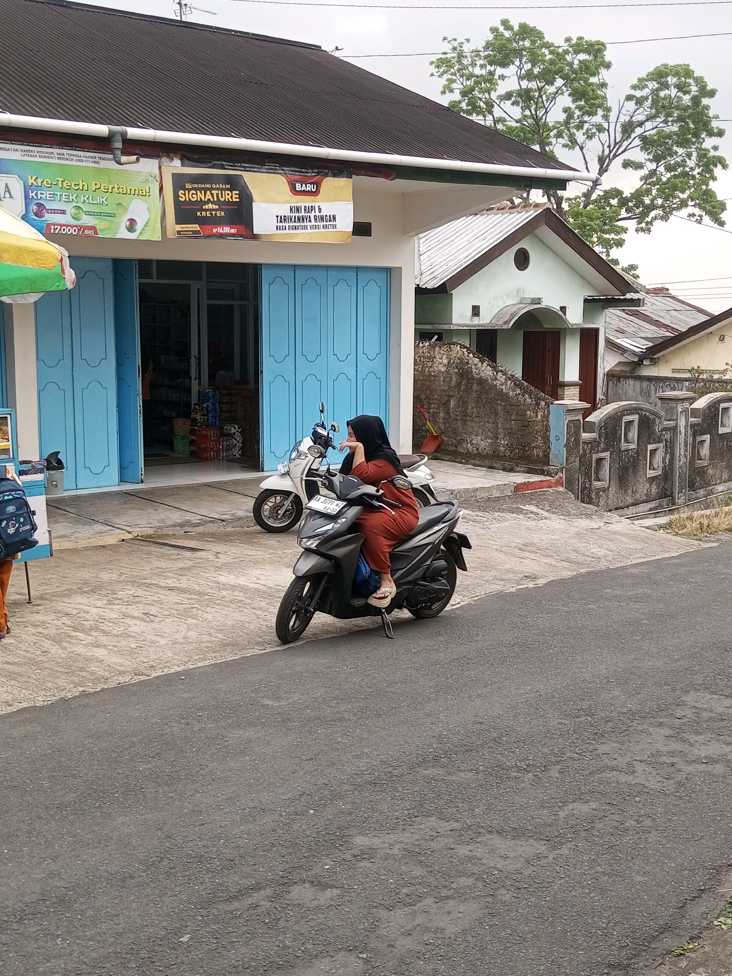 In Krasak, Indonesia, a woman wearing a black hijab and orange clothing is sitting on a dark gray scooter. She is leaning her head on her hand and looking to the right. The scooter is parked on a paved surface in front of a building with blue double doors and a dark roof. To the left of the scooter, a white scooter is parked. Above the building entrance, a banner advertises "Kre-Tech Pertama!" and "SIGNATURE KRETEK". The background shows residential buildings, trees, and power lines under an overcast sky. The scene appears to be a street in a semi-urban or rural area.