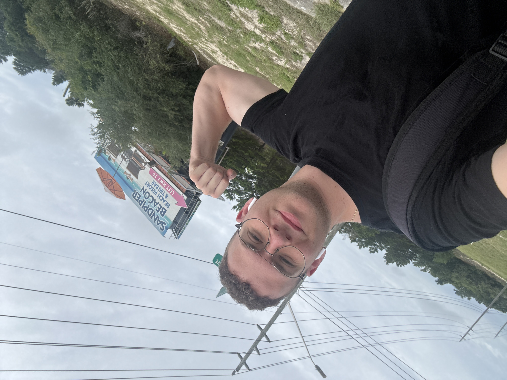 A selfie, taken at street level, features a man in a black T-shirt and glasses flexing his arm, with a "Sandpiper Beacon Beach Resort & Tiki Bar" billboard in the background. Power lines stretch across the sky above the street. The location is Panama City Beach, United States of America, based on the billboard and environment. A green street sign is present on a pole. There is greenery along the horizon.
