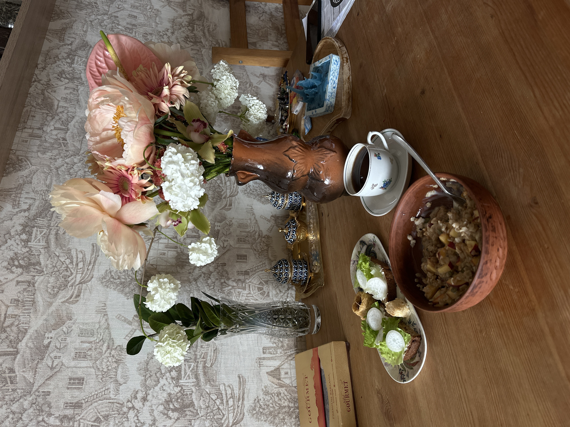 The scene depicts a tabletop still life, likely indoors. The table is split into two distinct surfaces: one covered with a patterned tablecloth featuring stylized buildings and the other, a plain wooden surface.
On the left, the table with the tablecloth is adorned with two vases containing flowers. One vase is brown and bulbous, holding an arrangement of pink peonies, anthuriums, and white hydrangeas. The second vase is tall, clear, and cylindrical, containing a smaller arrangement of white hydrangeas. Near the vases are decorative objects with blue and gold detailing and a box of gourmet baklava.
On the plain wooden surface to the right is a breakfast setting consisting of a bowl of oatmeal with fruit and a white teacup filled with a dark liquid. A spoon rests in the oatmeal bowl, suggesting it has been partially consumed. There is also a small plate containing a salad with what appear to be leafy greens, radish slices, and a small pastry item.
The location, Vladimir, Russia is inferred from the decorative objects which are in a style commonly associated with that region. There are no people visible in the scene. The main actions implied are the preparation and partial consumption of a meal.