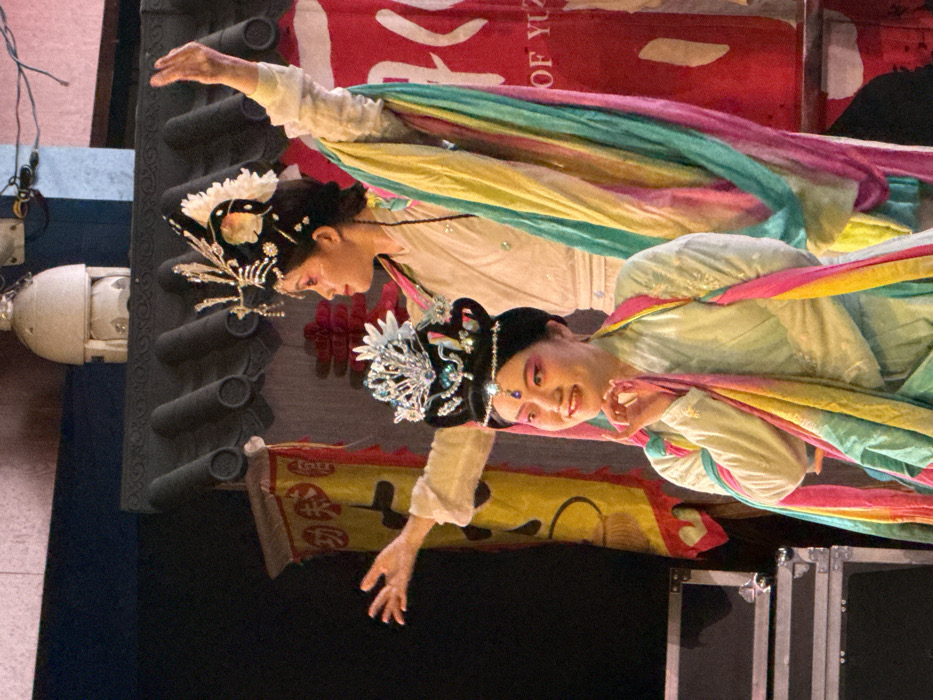 Two East Asian women in elaborate traditional Chinese performance attire are depicted on a stage in Yuzhong District, China. The woman in the foreground, positioned slightly left of center, faces right with her head turned towards the viewer, displaying a subtle smile. She wears a light green-blue long-sleeved garment and a silver and blue ornate headdress. Her face features prominent pink stage makeup around her eyes. Her right hand is gently raised towards her chin. The second woman stands behind and to the right, facing left in profile with her gaze directed slightly upwards. She wears a white or cream-colored long-sleeved garment and a black and gold ornate headdress with white floral accents. Her left arm is extended outwards, and her right arm is raised high above her head, both hands open. Both women have long, flowing multicolored sashes (pink, yellow, blue, green) draped over their arms and shoulders, indicating dynamic movement.
The background includes stage elements: a dark grey structure resembling stylized roof eaves across the upper middle, and several banners. To the left, a yellow banner with red Chinese characters and a fringed edge is partially visible. To the right, a large red banner with white Chinese characters is visible, prominently displaying the English text "OF YUZ", indicating the location. A white dome-shaped surveillance camera or stage light fixture is mounted high on the back wall. On the bottom left, several stacked black equipment cases are visible. The scene is brightly lit, focusing on the performers, against a largely dark and indistinct background elsewhere.