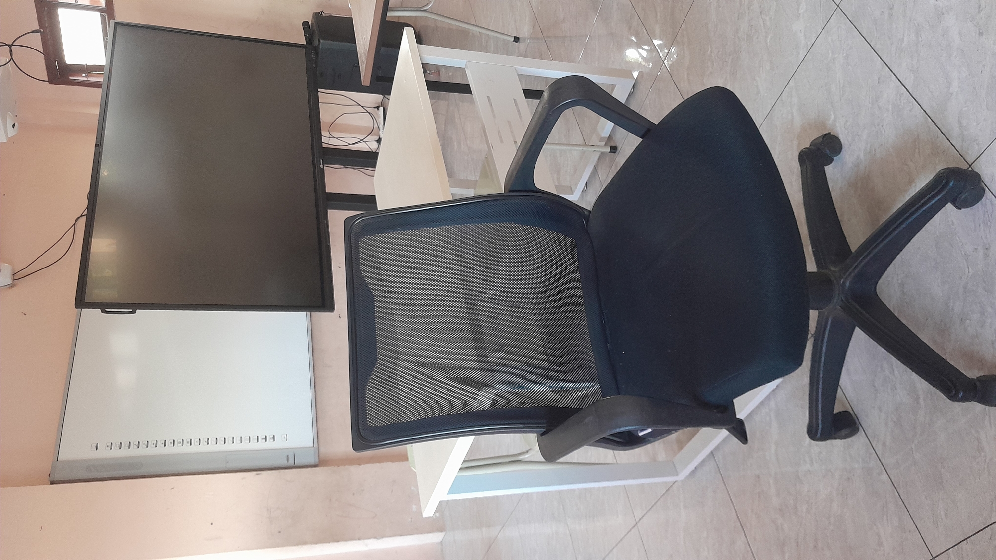 A black mesh office chair with armrests and a wheeled base is positioned on a tiled floor in front of a white table. Behind the table, a large, dark interactive display screen is mounted on a stand, and to its left, a whiteboard with markings is affixed to the wall. The environment appears to be a classroom or meeting room, with light pink walls and a window visible in the background. The scene is lit by ambient light. No people are present, and no actions or interactions are depicted.