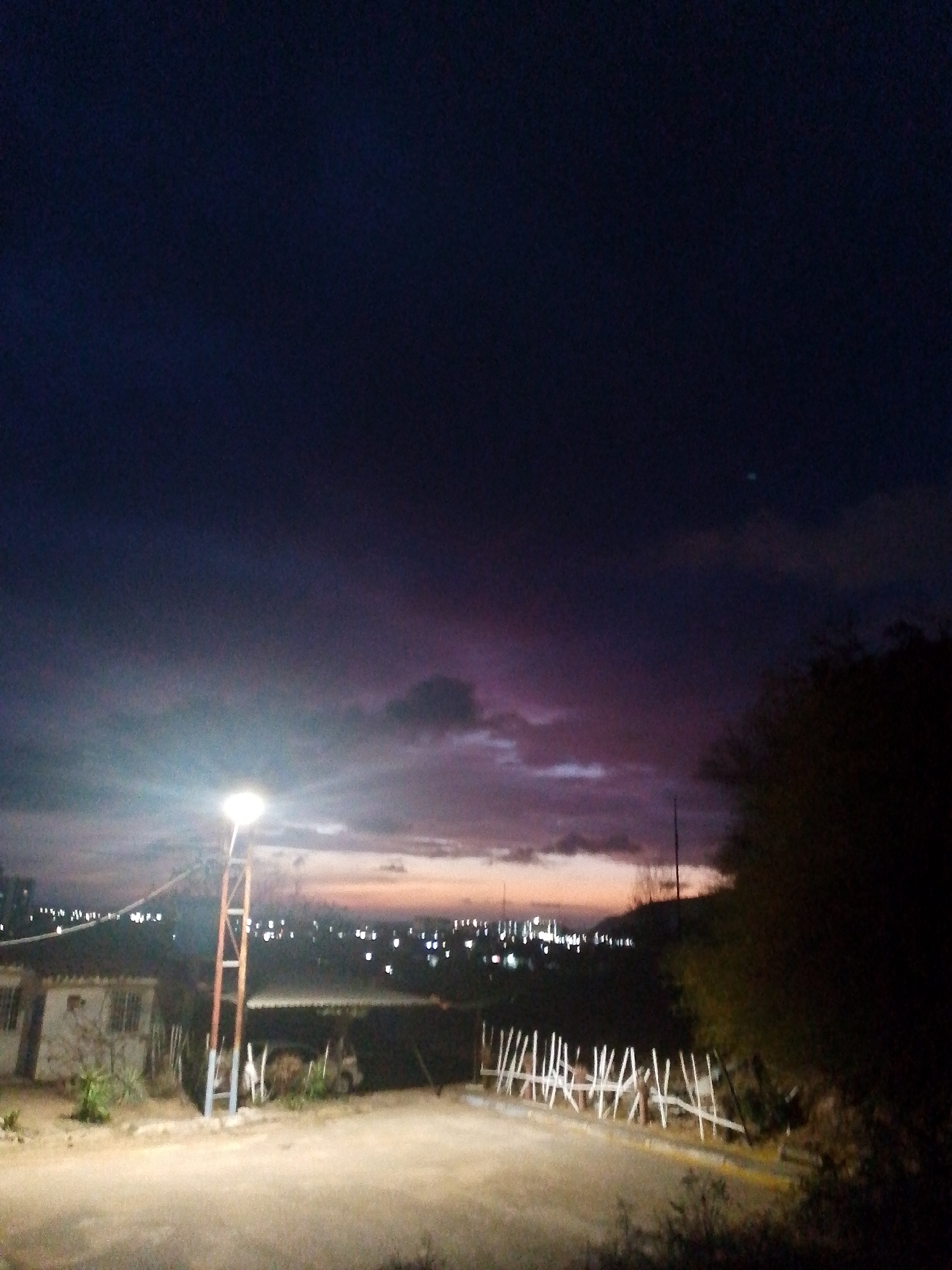 At night in Juan Griego, Venezuela, a street is illuminated by a streetlight. A building is to the left of the streetlight, and a fence is on the right. In the background, city lights are visible. Dark sky above.