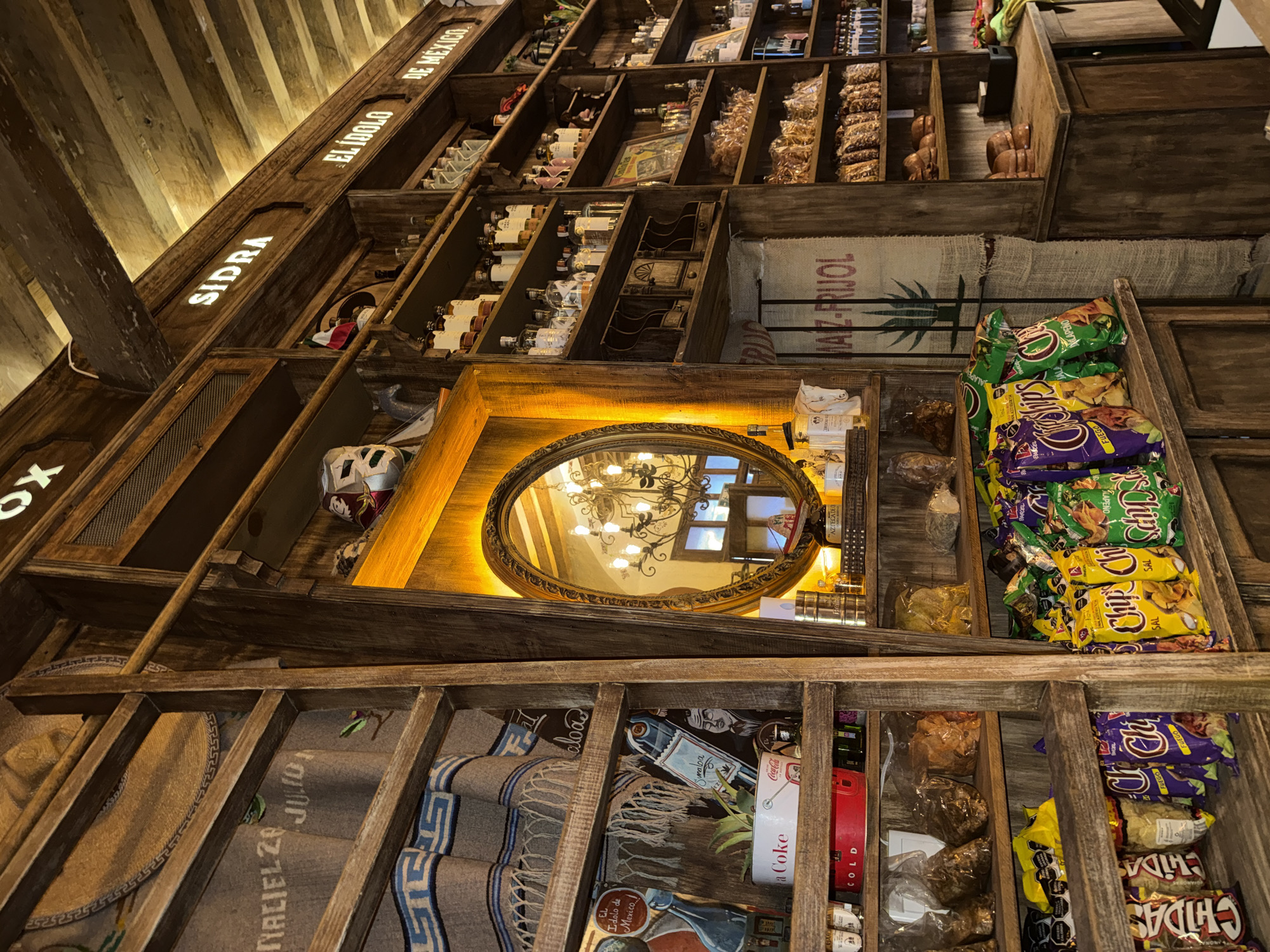The image captures a view of shelves displaying various products, possibly within a store located in Puebla, Mexico, as indicated by the "El Idolo de Mexico" signage. The shelves are constructed from wood and house an assortment of items, including bottles of liquor (presumably Sidra), bags of chips, hats, blankets, and what appear to be traditional Mexican snacks and goods. A notable feature is a decorative oval mirror positioned among the shelves. The setting suggests an interior space, likely a store or market, with the displayed items implying a retail or commercial function. The lighting is warm, possibly from overhead fixtures, creating a cozy atmosphere. The arrangement of items on the shelves reflects a mix of commercial products and cultural artifacts, indicative of a store catering to tourists or locals interested in regional specialties.