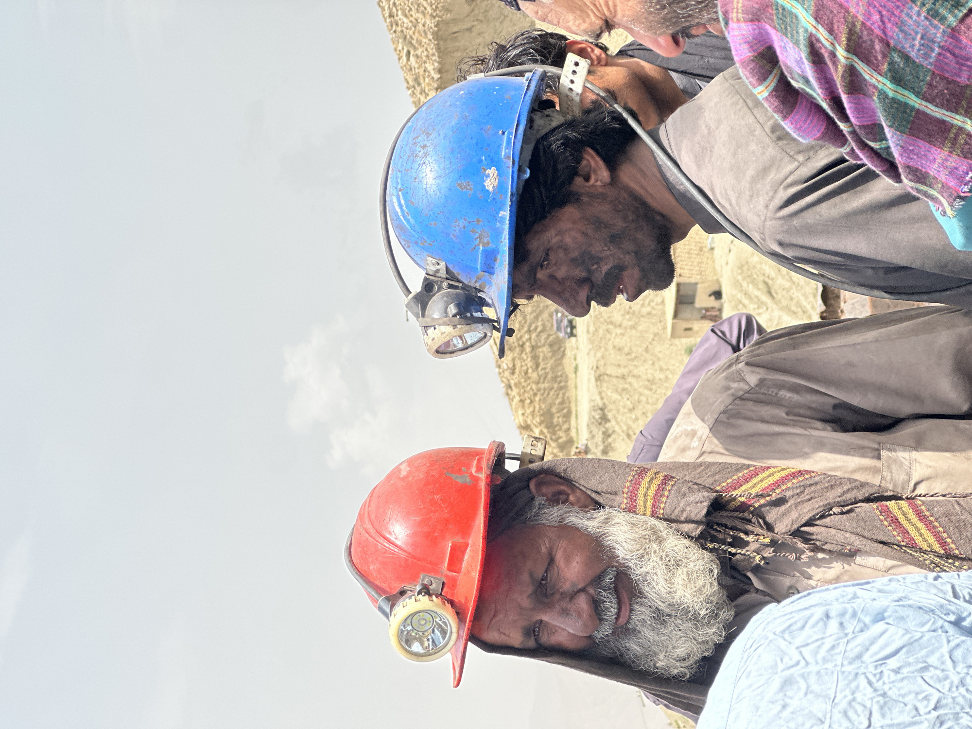 The image depicts a group of men in what appears to be a mining or quarrying area, likely near Quetta, Pakistan, given the dusty, arid landscape of hills and sparse vegetation. Two men in the foreground are prominently featured, both wearing hard hats with headlamps, suggesting they are engaged in work that requires protection and illumination.
The man on the left has a red hard hat, a long gray beard, and is wearing a brown scarf with red and yellow stripes around his neck. His face is smudged with dirt. He is looking slightly to his left.
To the right of him, another man wears a blue hard hat and his face is also significantly covered in dirt, particularly around his mouth and chin. He is looking to his right. Behind these two men, other individuals are partially visible, one wearing a plaid shirt with purple, green, and pink colors. In the background, there are dusty hills and a small building. A vehicle can be seen in the distance. The sky is overcast with light clouds. The overall impression is of a working environment in a dry, possibly remote, location.