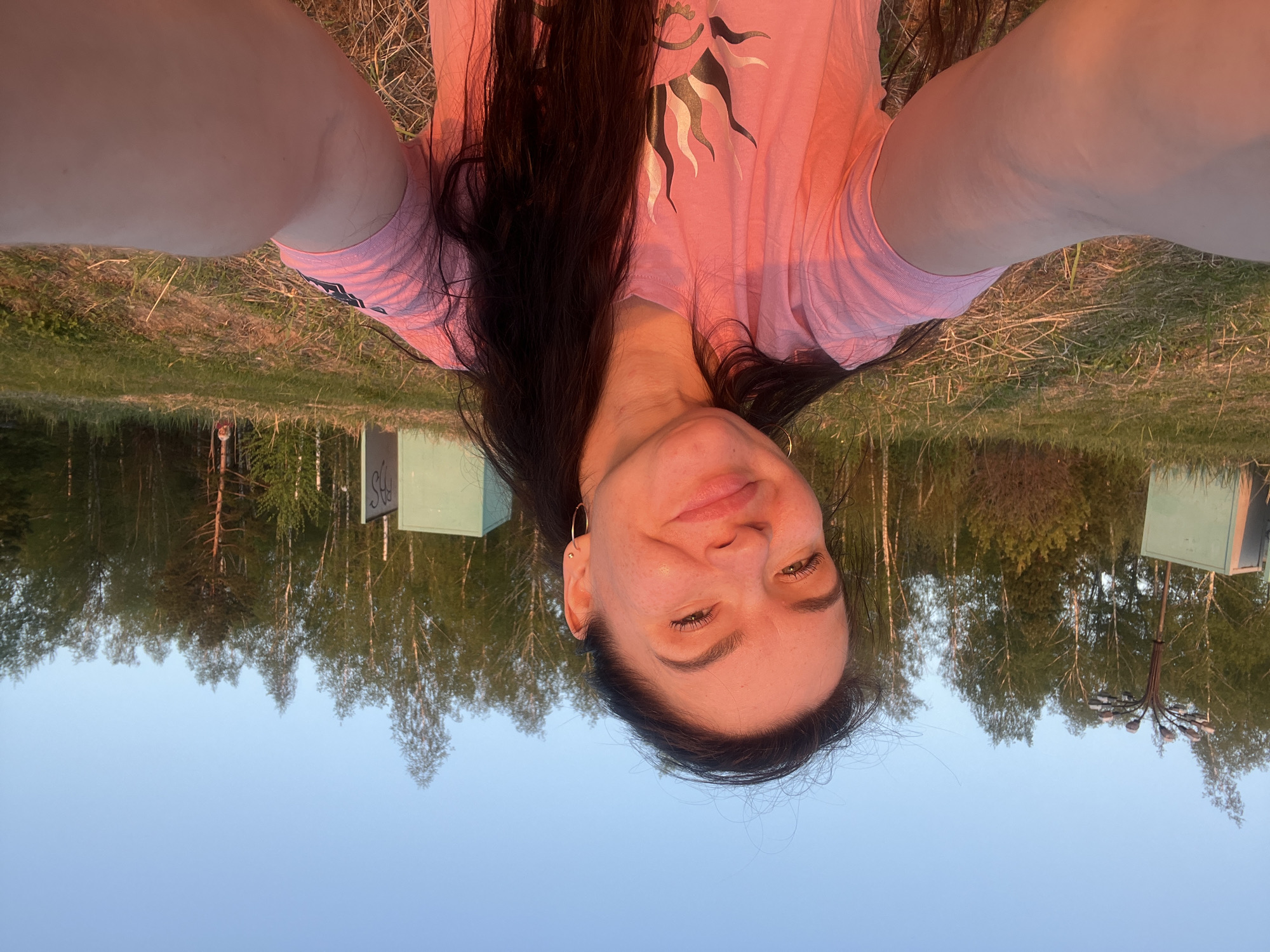 The image shows a woman taking a selfie in Borovoye Matyushino, Russia. She is wearing a pink t-shirt with a sun design. The background includes trees, grass, and what appears to be street lighting or signs. The sky is visible above the trees.